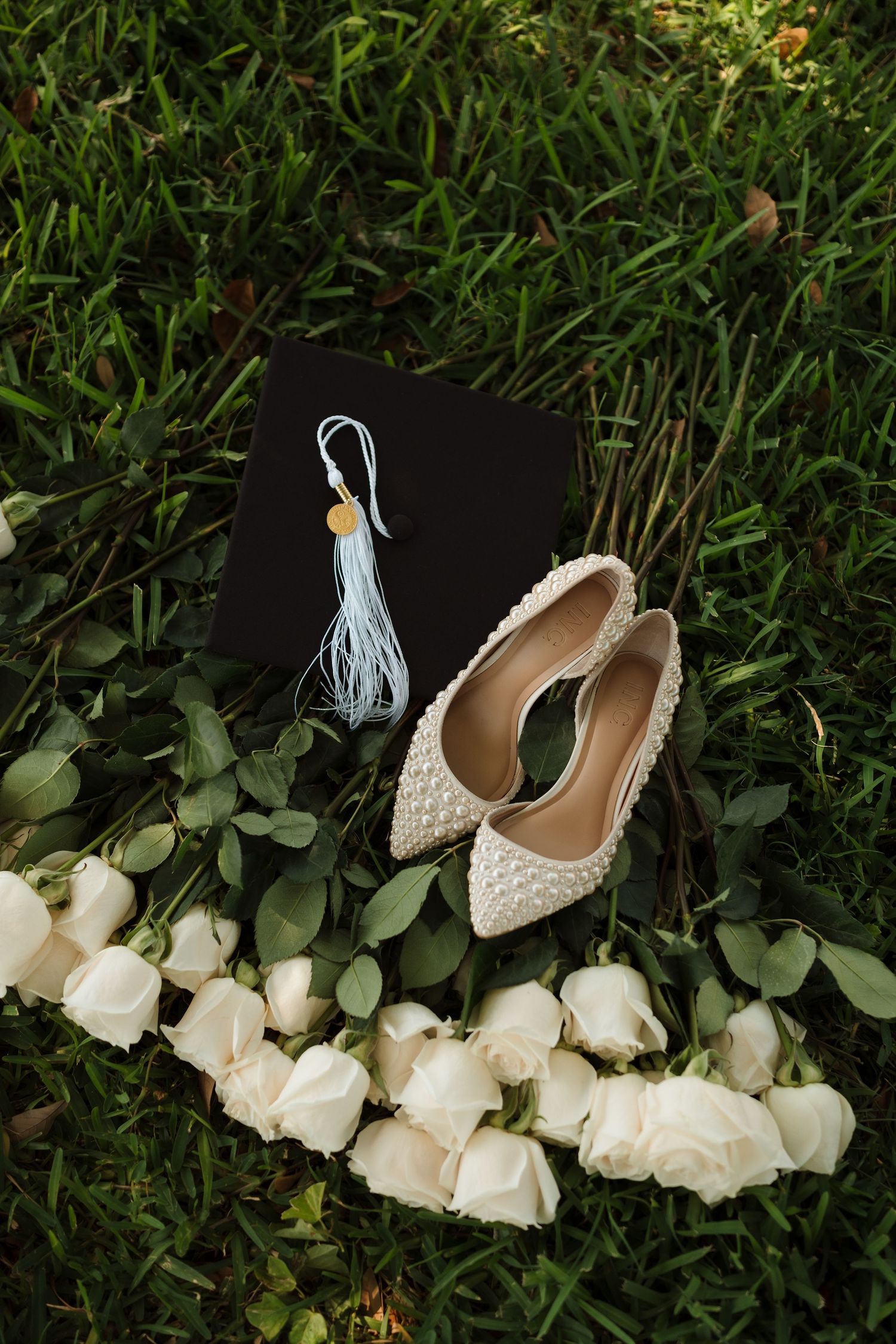 Elegant white wedding shoes with pearl details arranged with white roses on green grass.