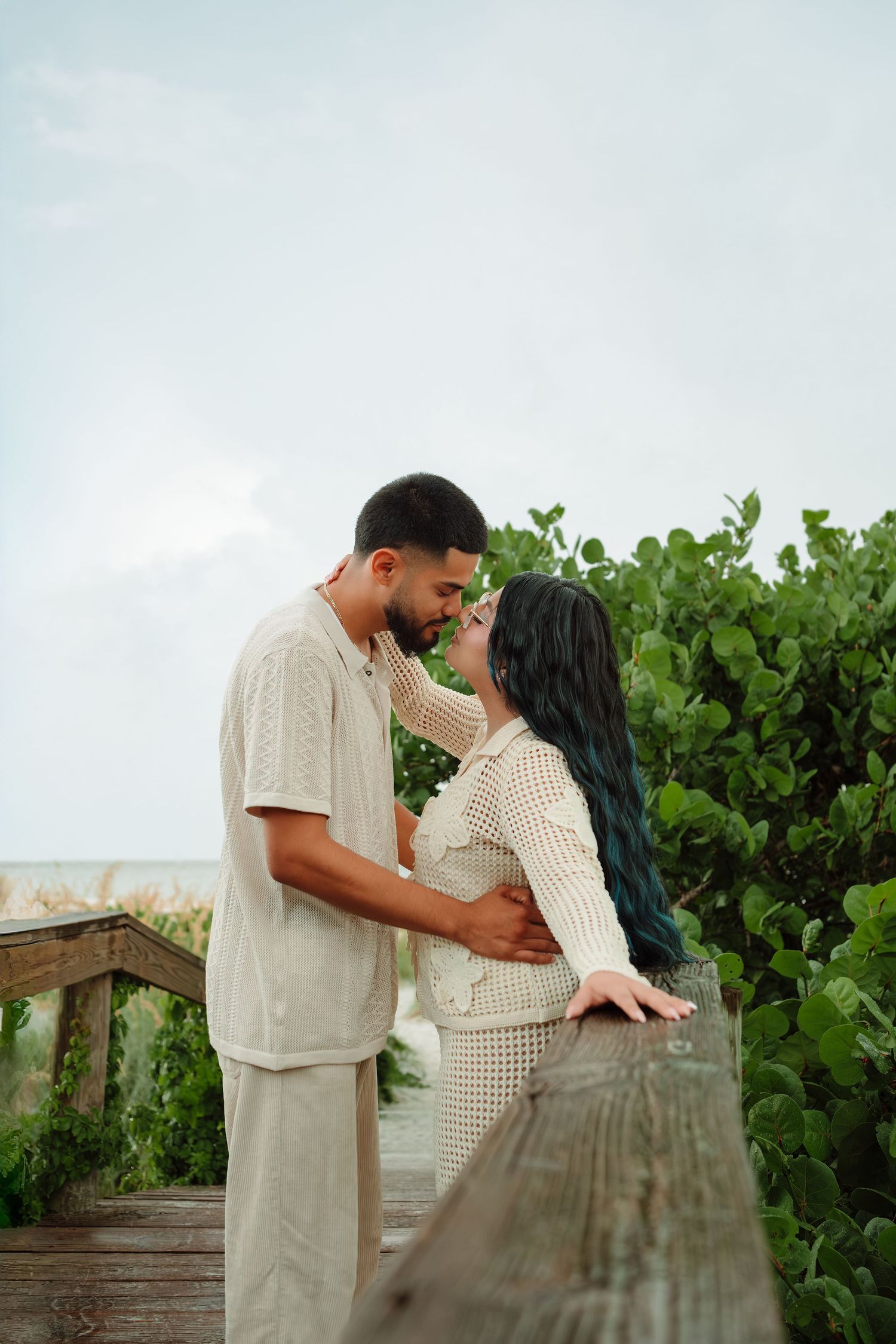 A couple shares a romantic moment on a beachside boardwalk, surrounded by lush greenery, dressed in matching cream outfits.