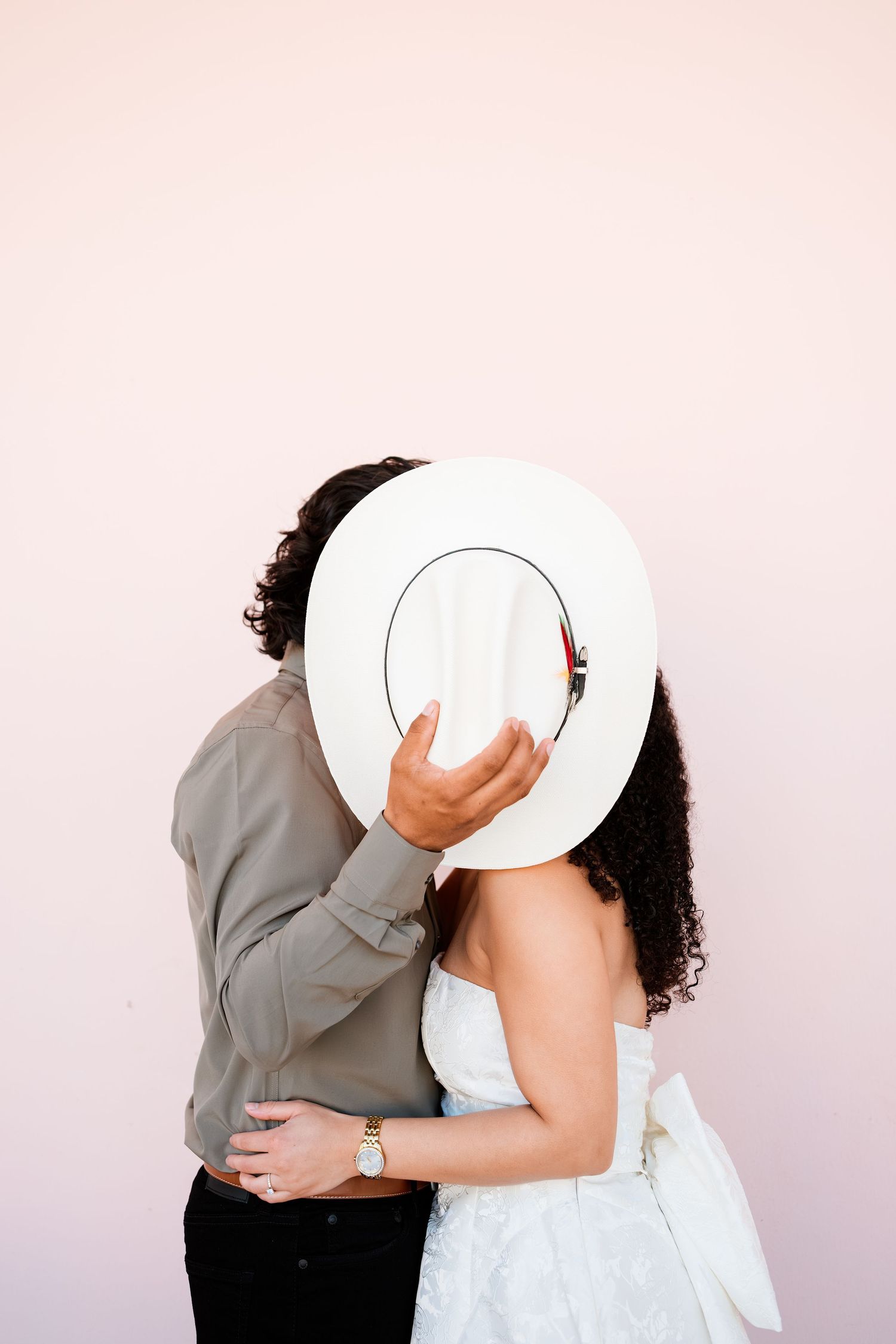 A couple sharing a kiss hidden behind a white wide-brim hat against a soft pink wall.