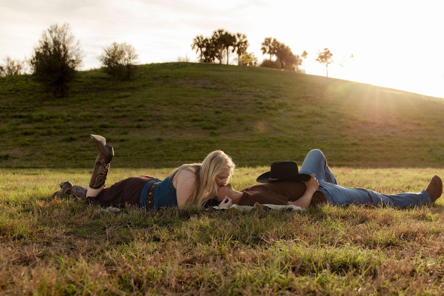 Couple lying in a sunny green field at golden hour, woman in jeans and man wearing a cowboy hat.
