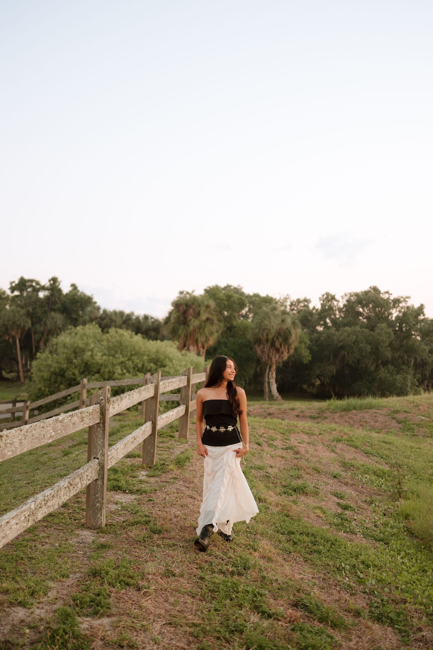 Woman in black top and white skirt walking along a wooden fence on a green rural path at dusk.