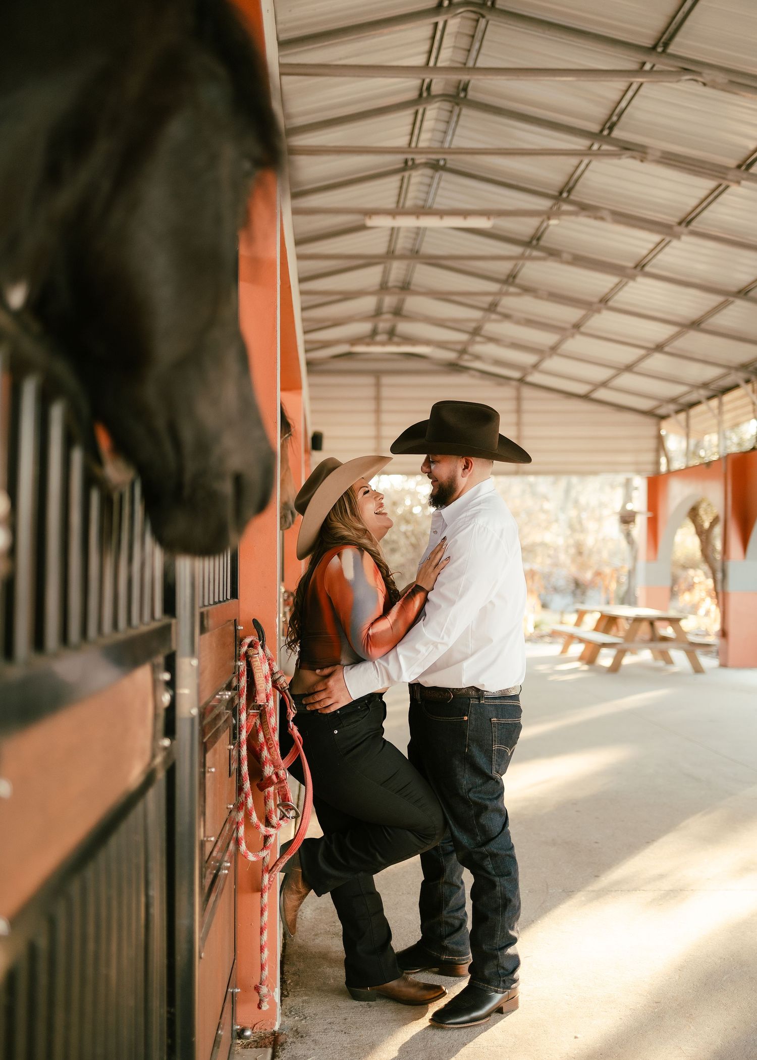 Couple in cowboy hats embrace romantically in a rustic horse stable, wearing western attire with boots and jeans.