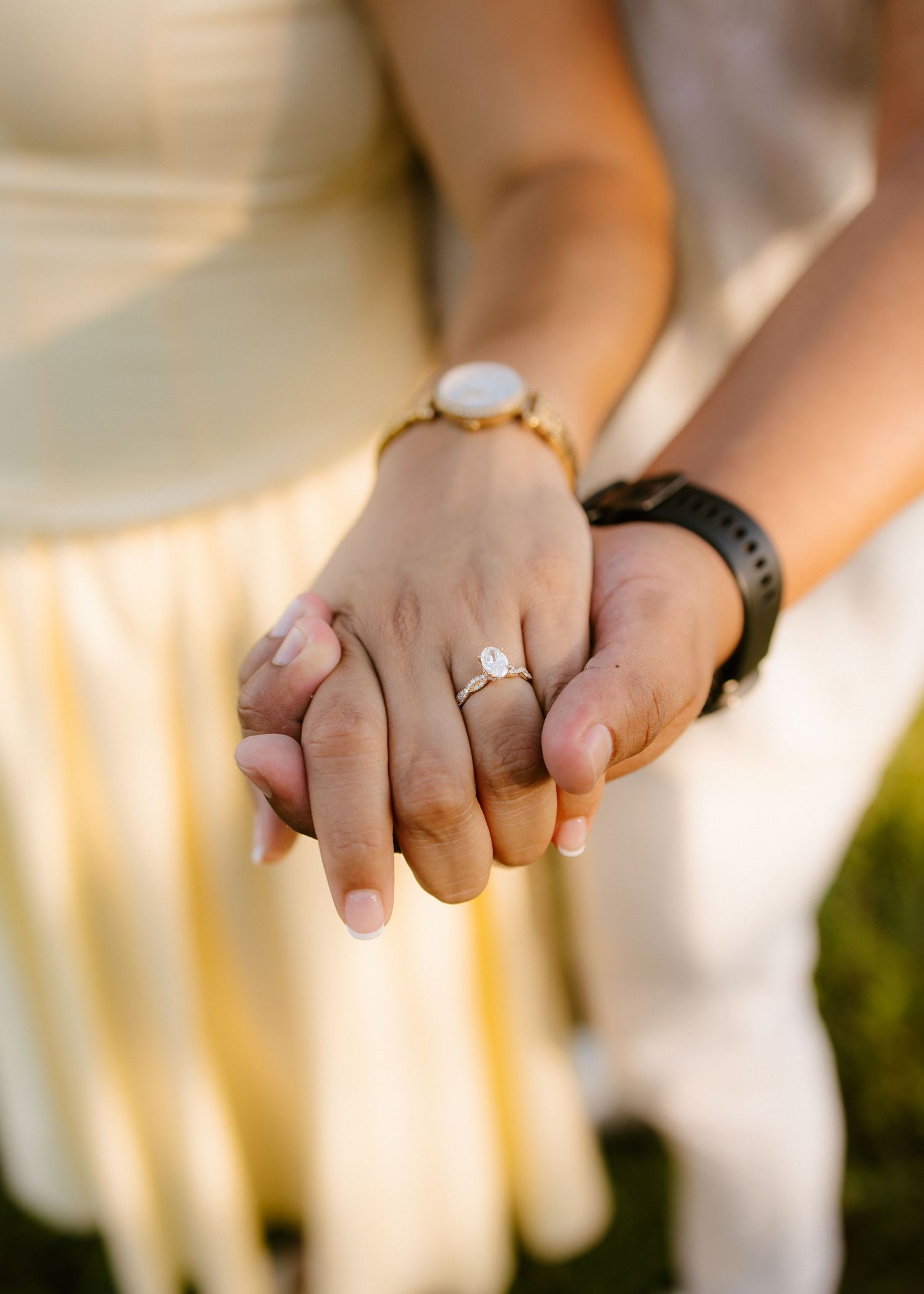 Couple holding hands showing engagement ring and watches in warm golden light.