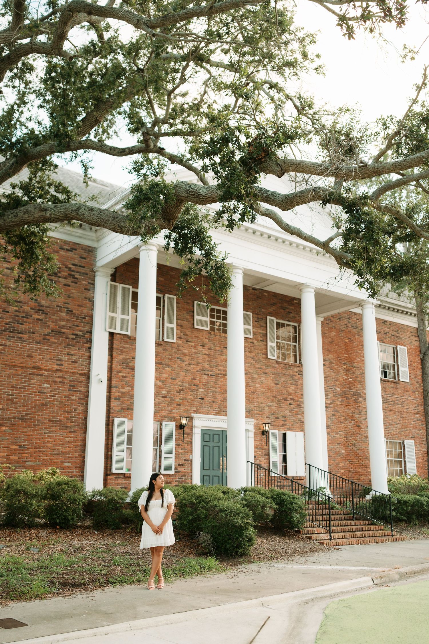 Woman in white dress standing before a grand brick building with white columns and large oak trees.