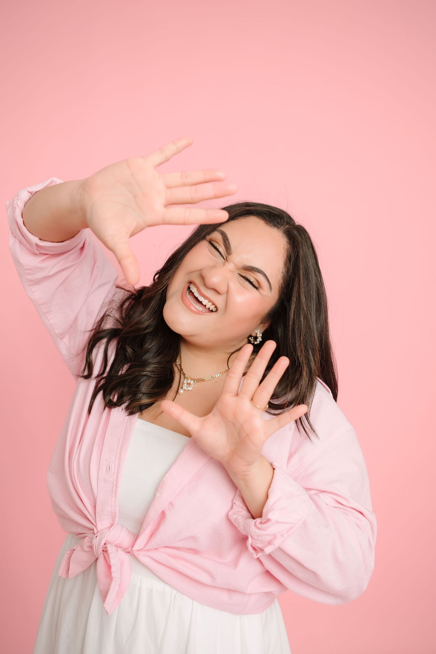 Smiling brunette woman in pink shirt making a frame gesture with hands against a pink background.