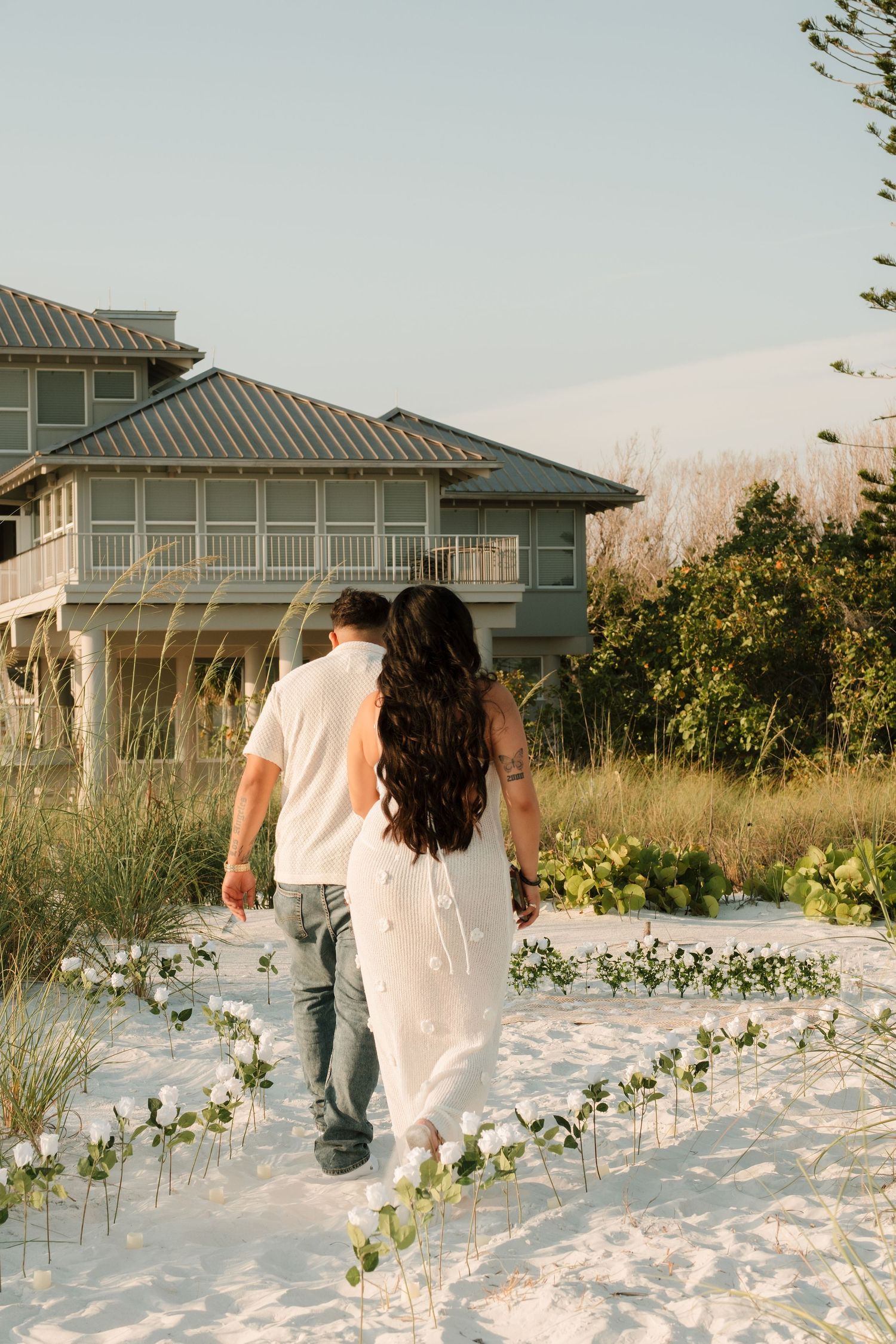 Couple walks hand-in-hand on white sand beach toward coastal home, surrounded by a floral heart arrangement at golden hour.