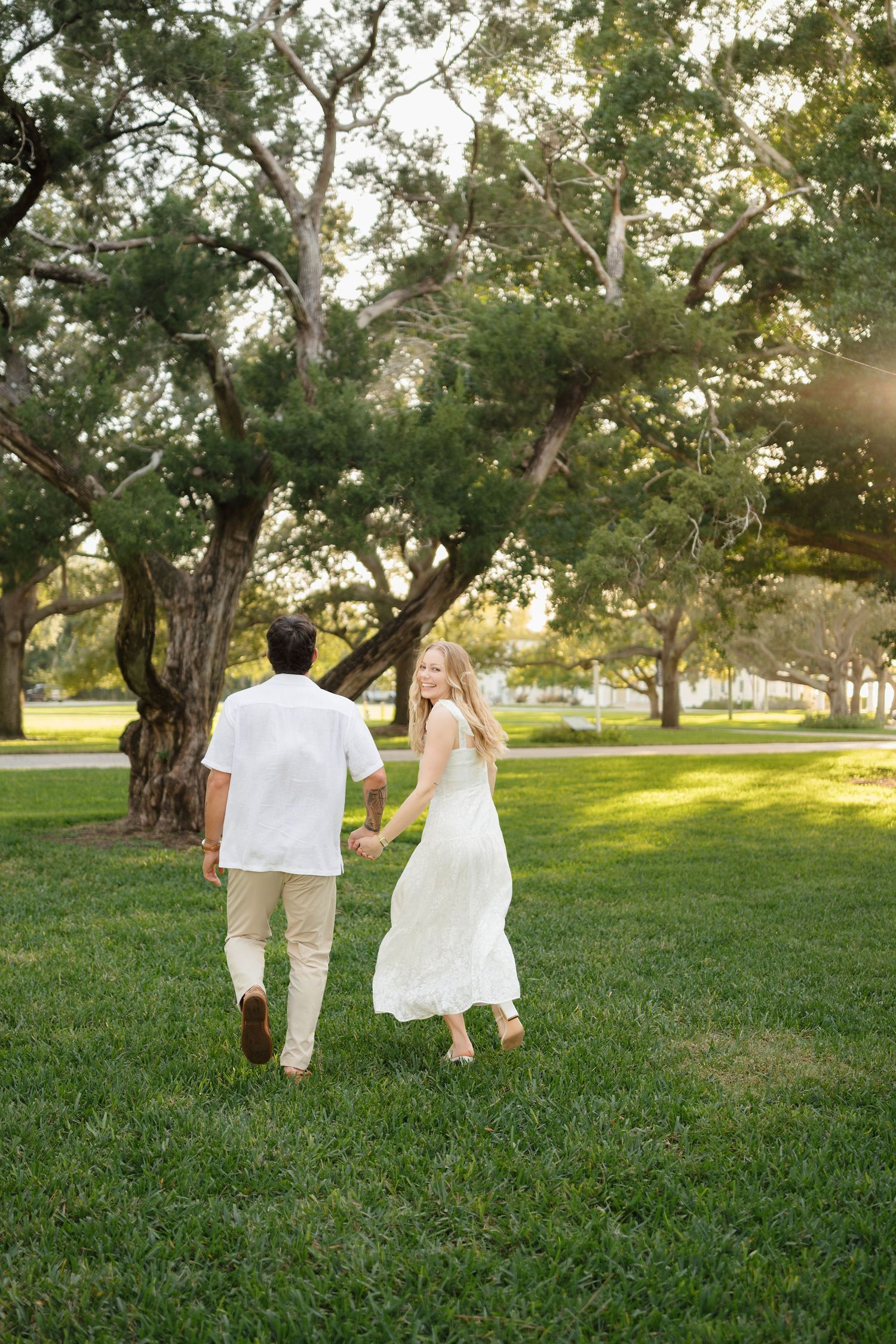 Couple holding hands walking through a sunny green park, woman in white dress glancing back, surrounded by large trees.