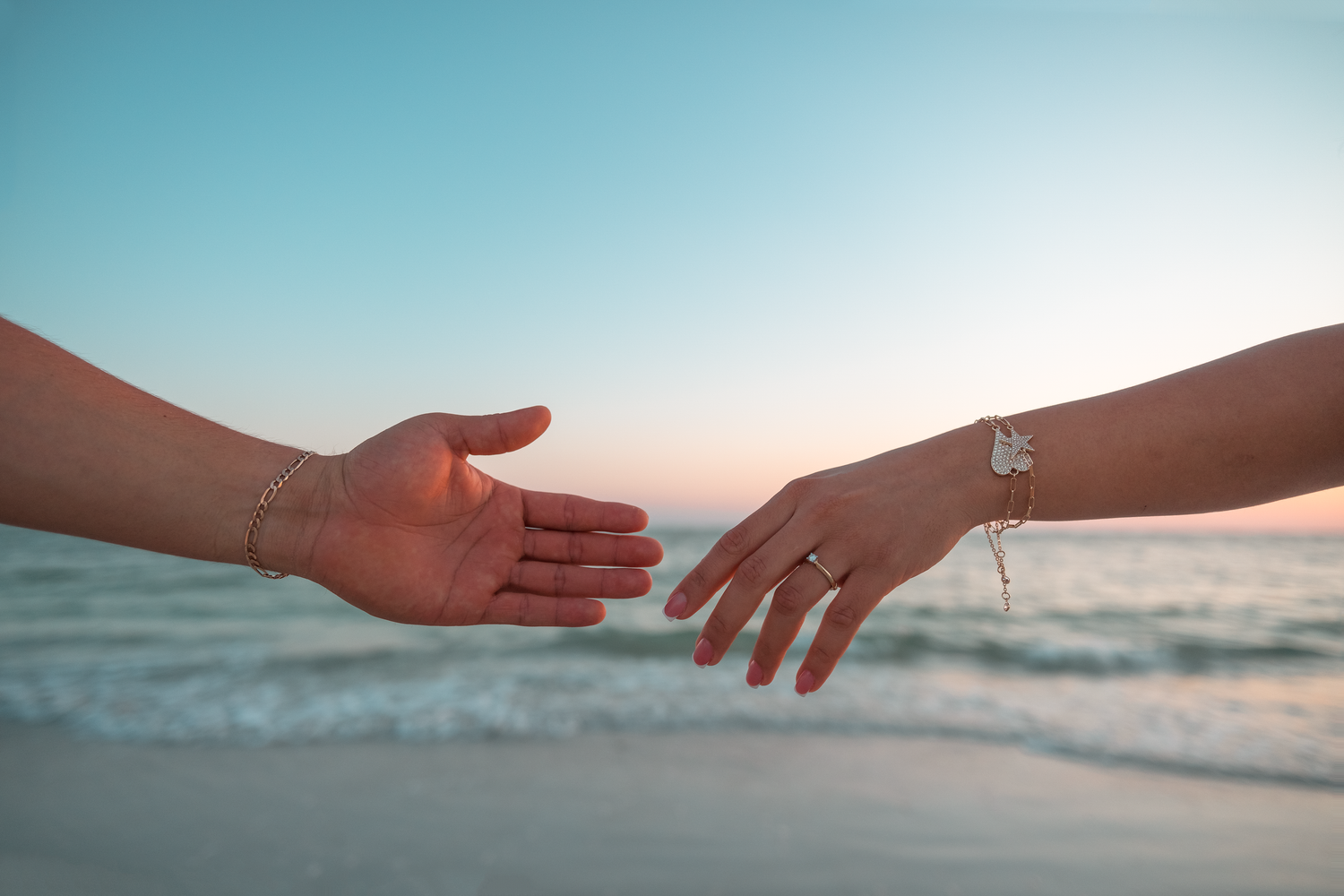 Two hands reaching toward each other on a beach at sunset, one wearing a bracelet and ring.