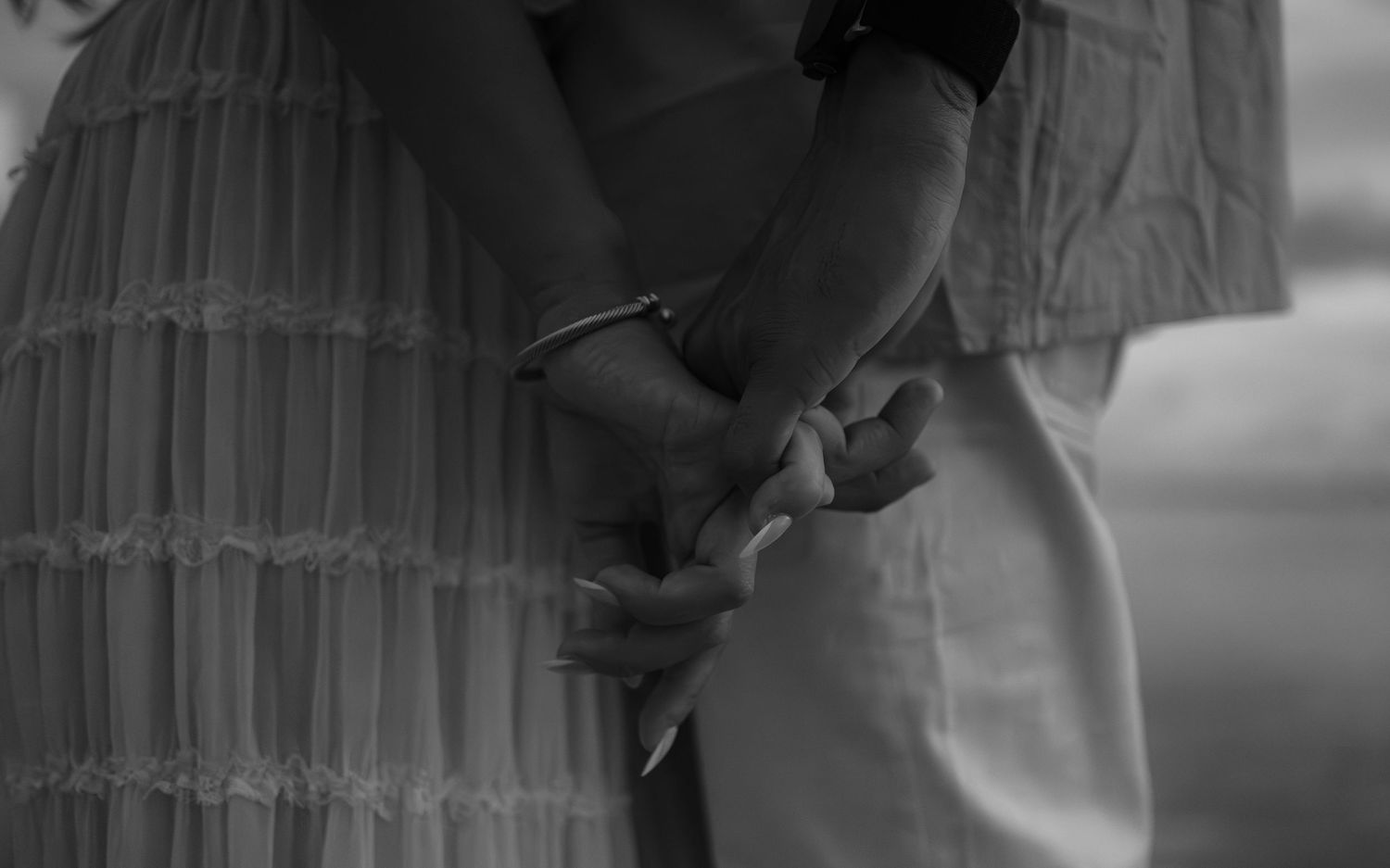 Black and white photo of two people holding hands tenderly, wearing light dresses outdoors.