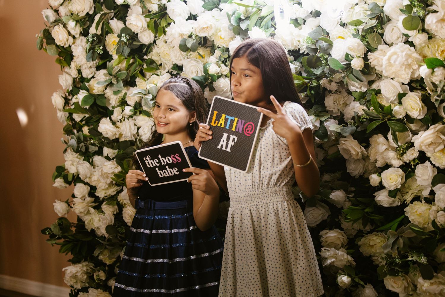 Two young girls posing with fun photo booth signs in front of a white floral wall at a celebration.
