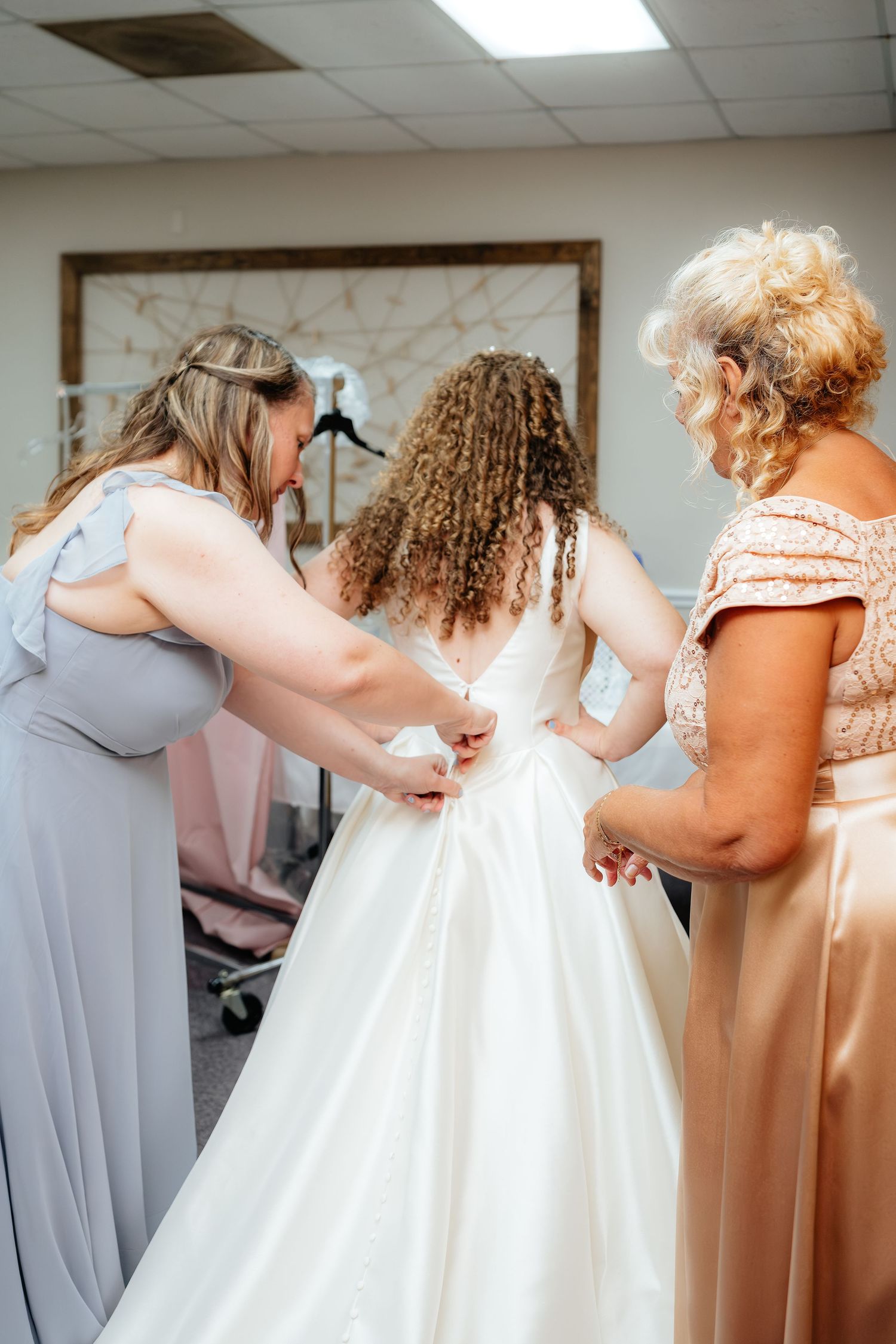 Two women help a bride button her white wedding gown in a bridal boutique fitting room.
