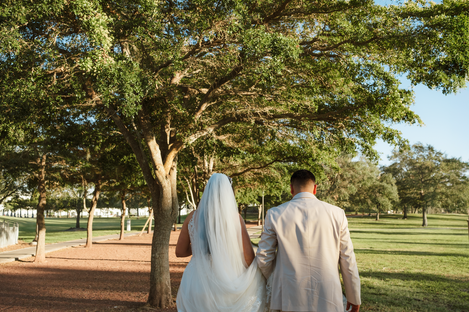 Bride and groom walking hand in hand under lush green trees in a sunlit park on their wedding day.
