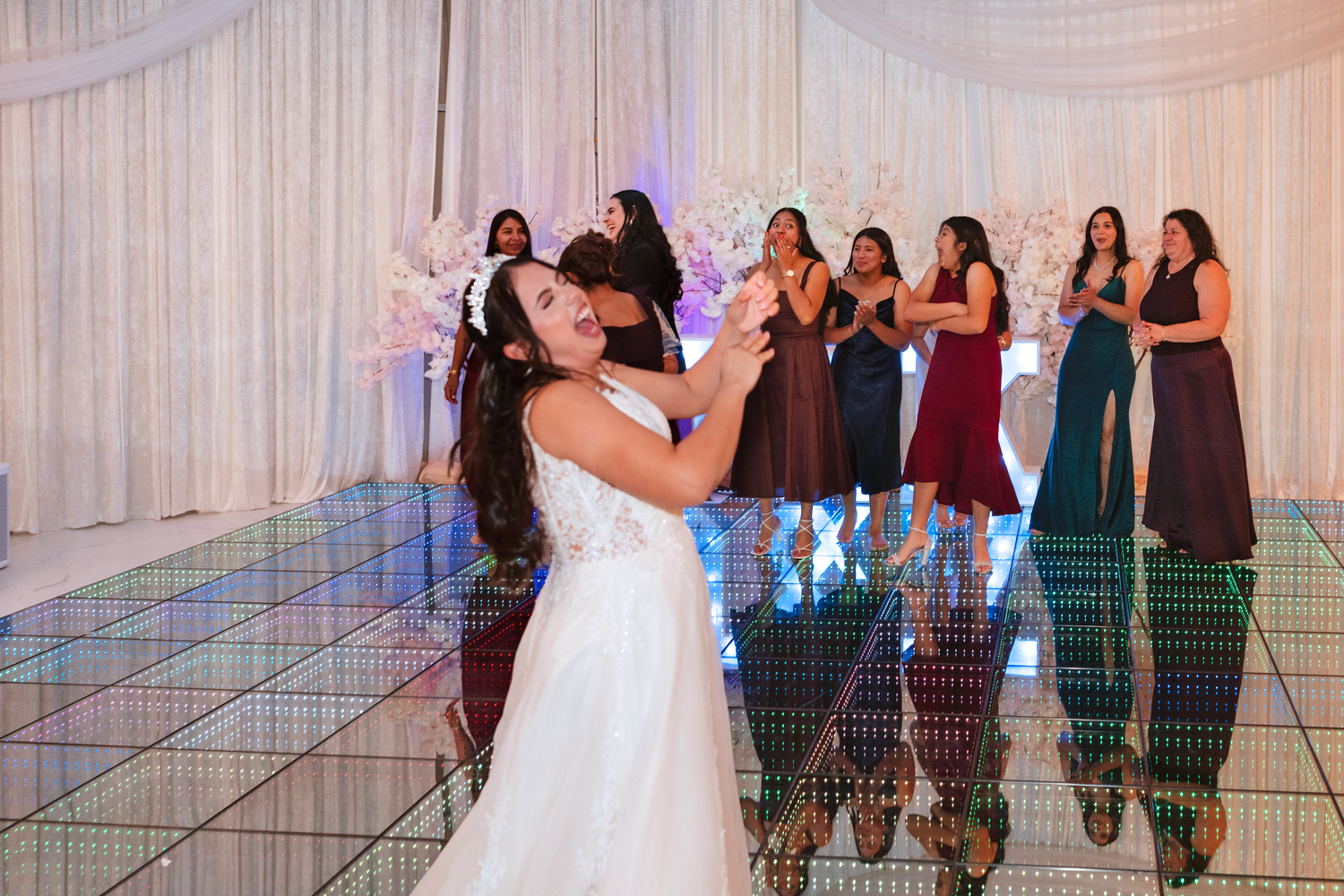 Bride tosses bouquet to excited bridesmaids in colorful dresses on mirrored dance floor.