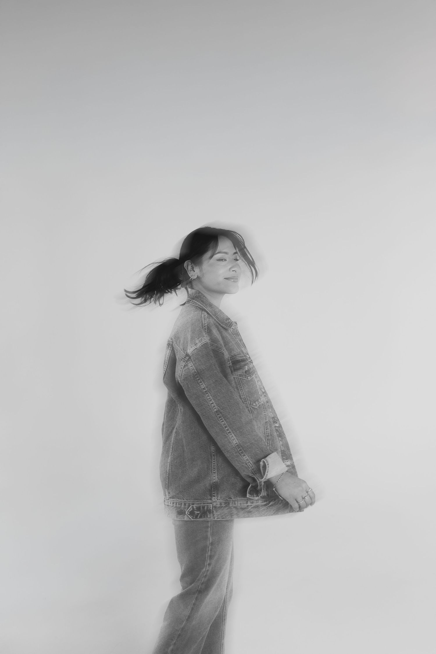 Black and white artistic portrait of a person in denim jacket and jeans turning with hair in motion against plain background.