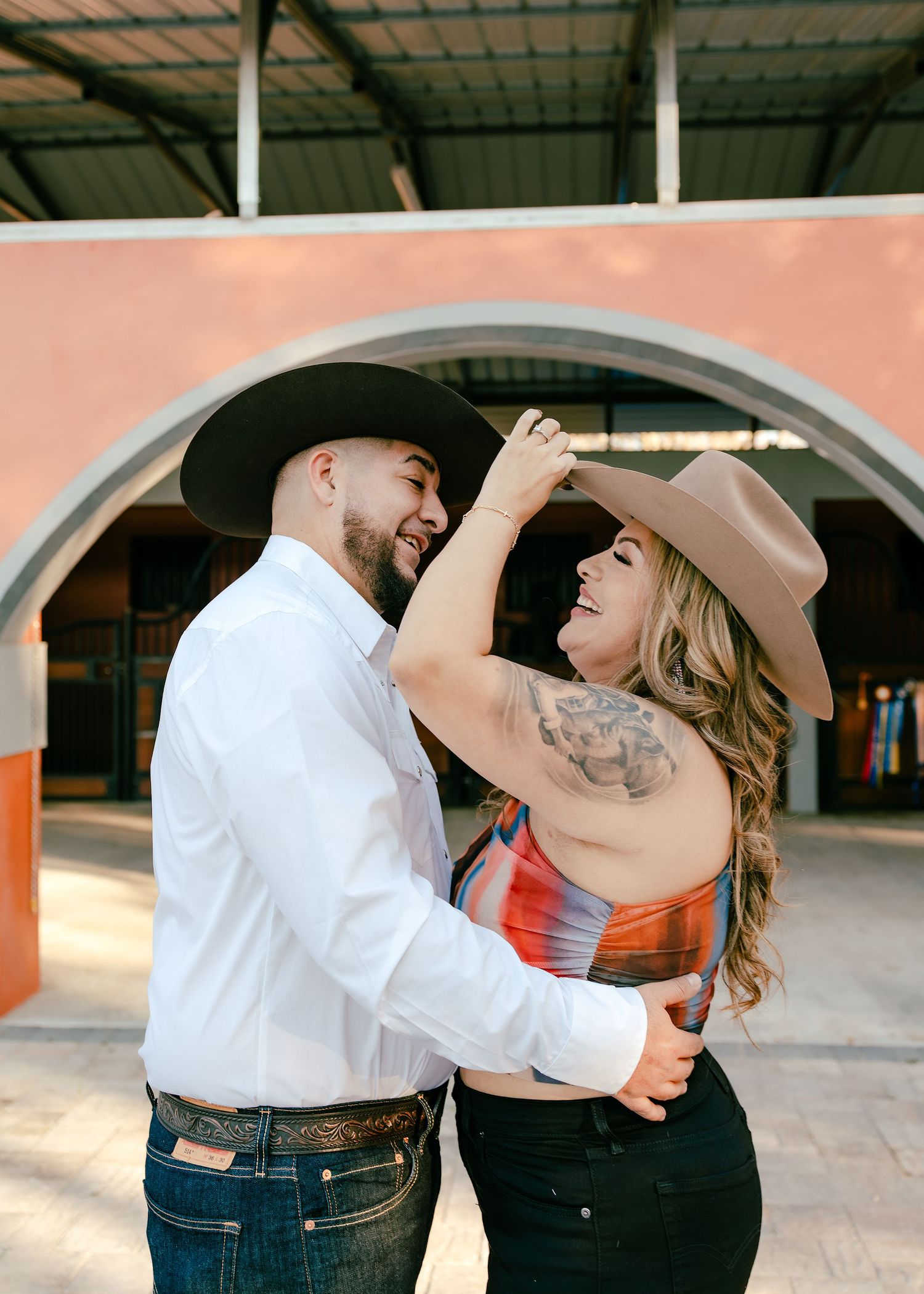 Couple in cowboy hats dancing and laughing outdoors near a colorful arched building.