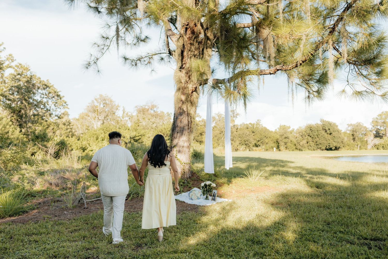 Couple holding hands walking toward a tree decorated with flowers in a sunny green park setting.