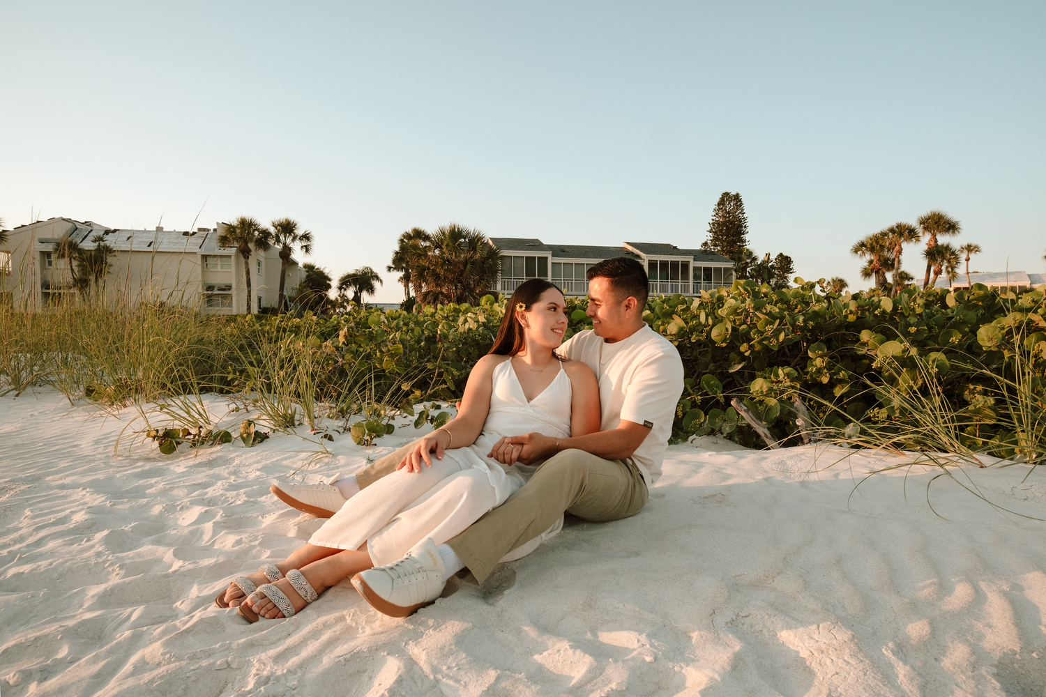 Couple sitting together on white sand beach at sunset with tropical greenery and beach houses in background.