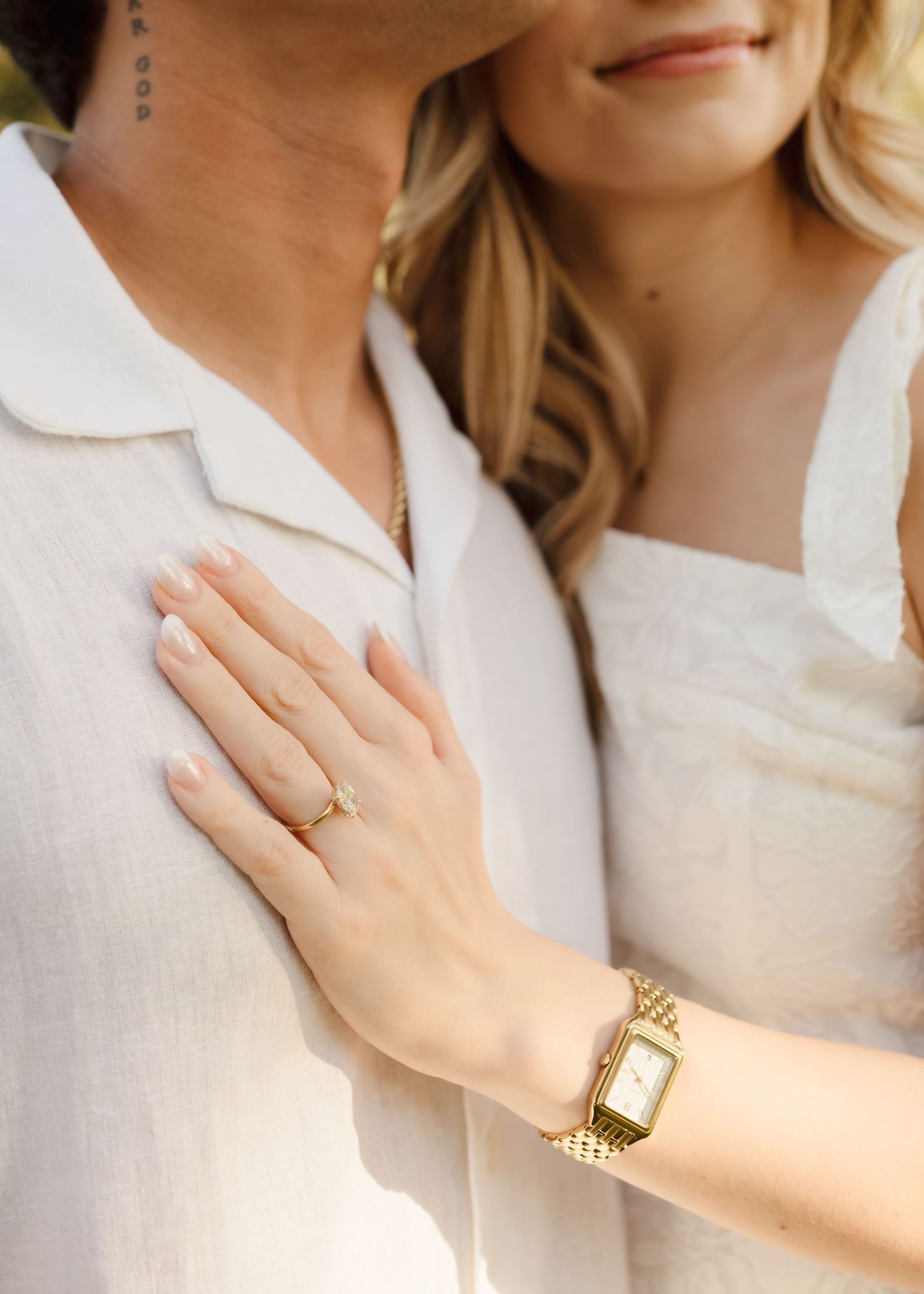 Woman in white dress with gold watch and ring rests hand on mans white linen shirt during engagement session.