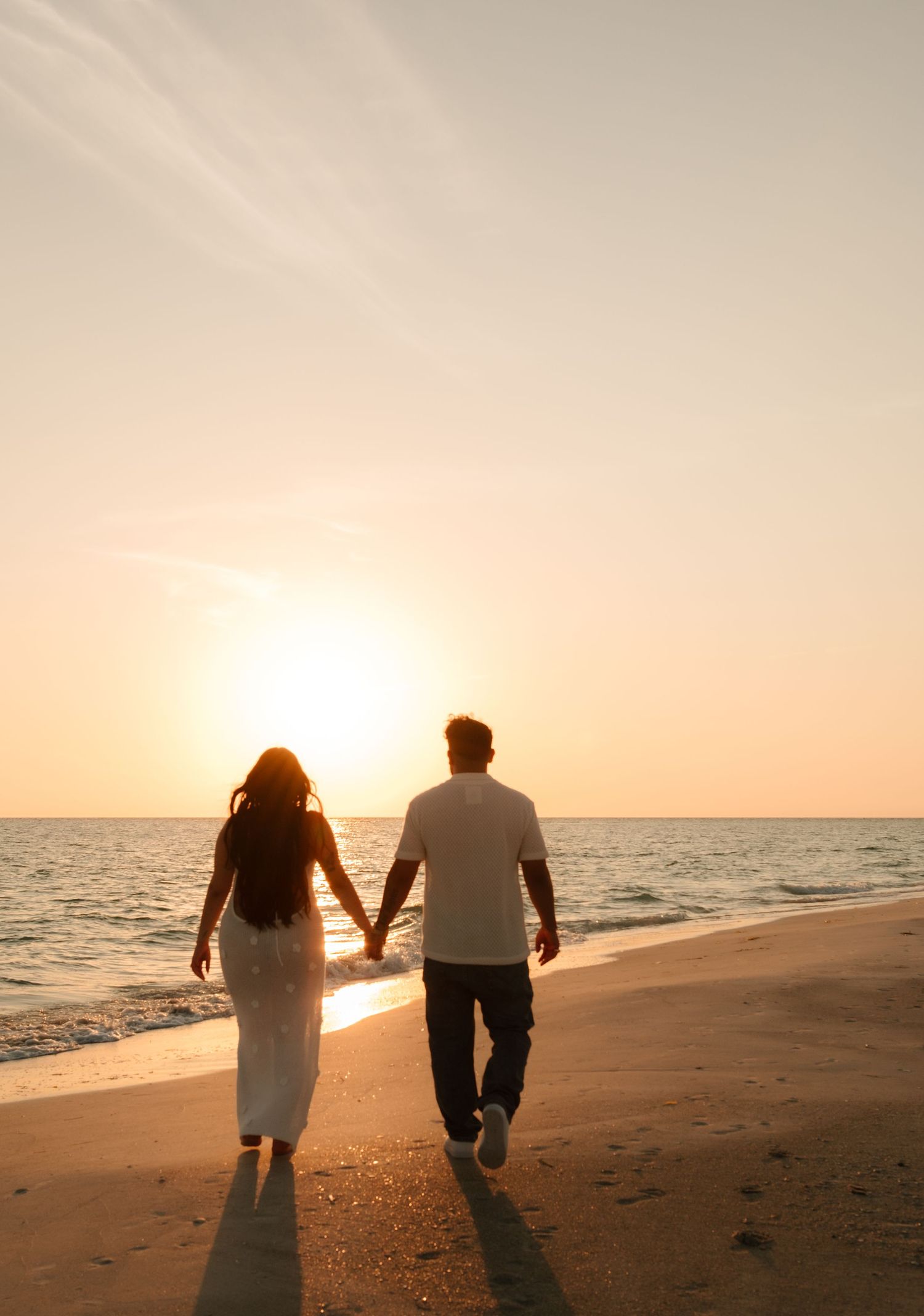 Couple holding hands walking on a sandy beach at golden sunset, silhouetted against warm orange sky and calm ocean waves.