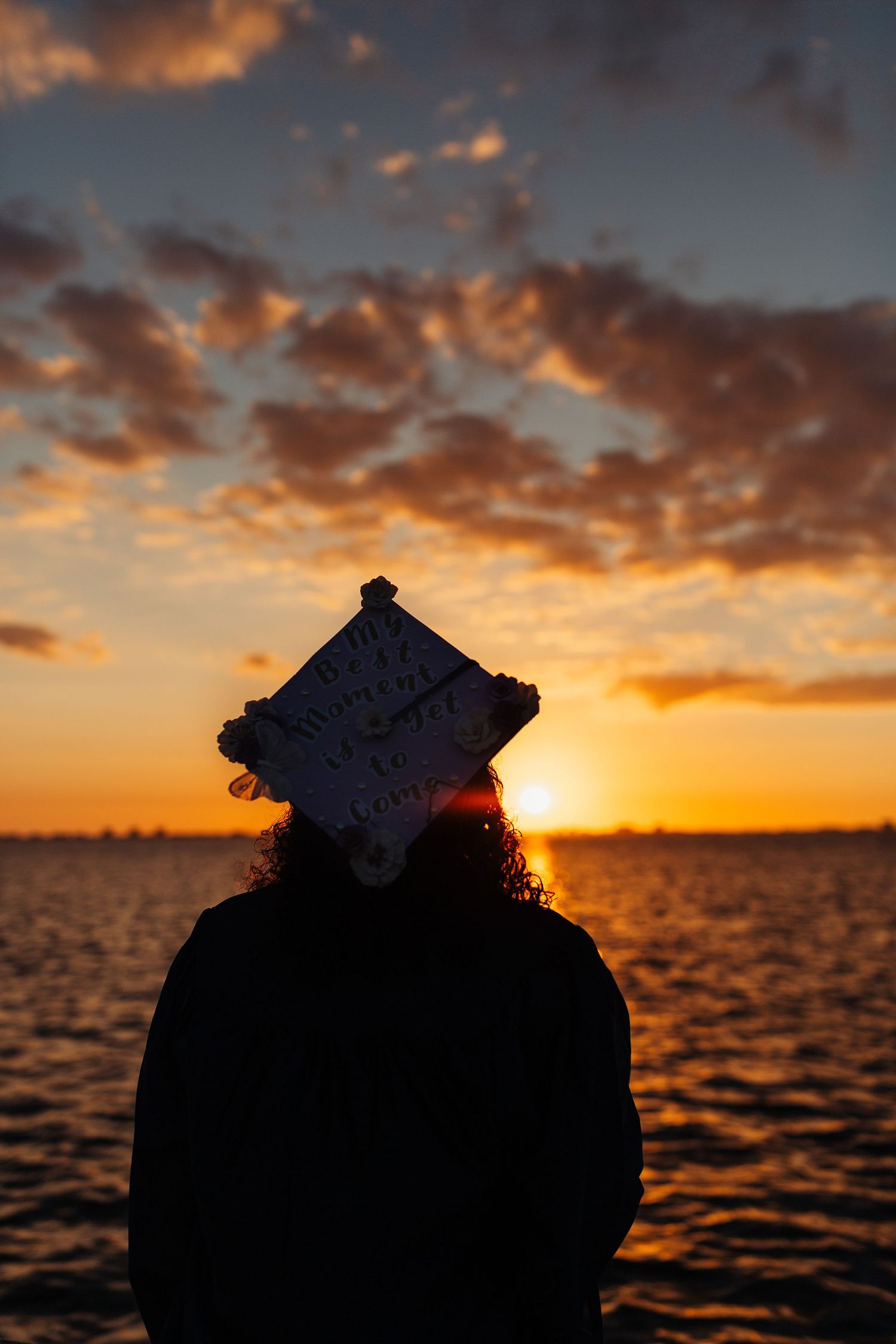 Silhouette of a graduate wearing a decorated cap against a vibrant orange sunset over the ocean.