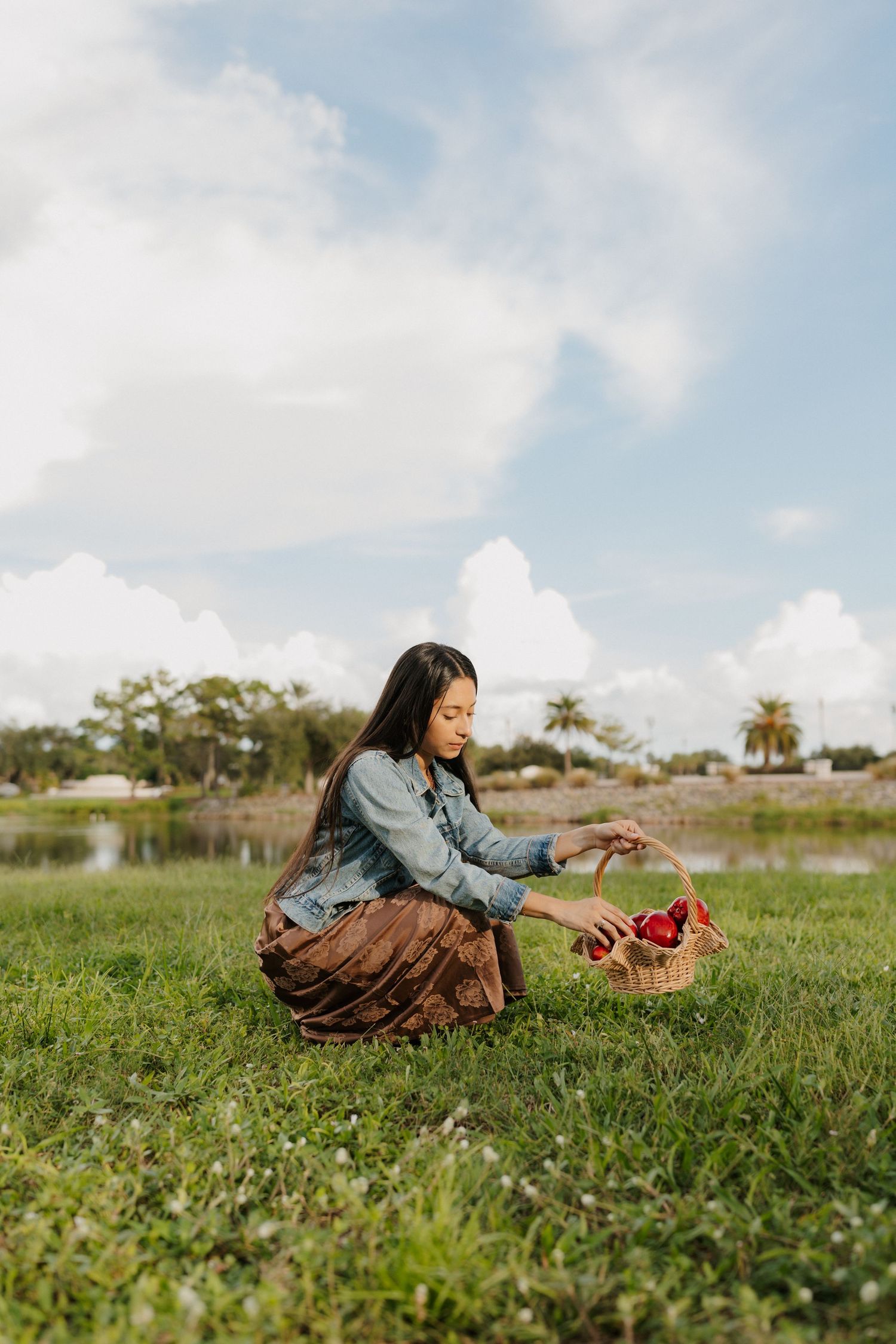 Woman in a denim jacket and brown skirt crouches on a grassy field by a pond, picking red apples into a woven basket under a cloudy sky.