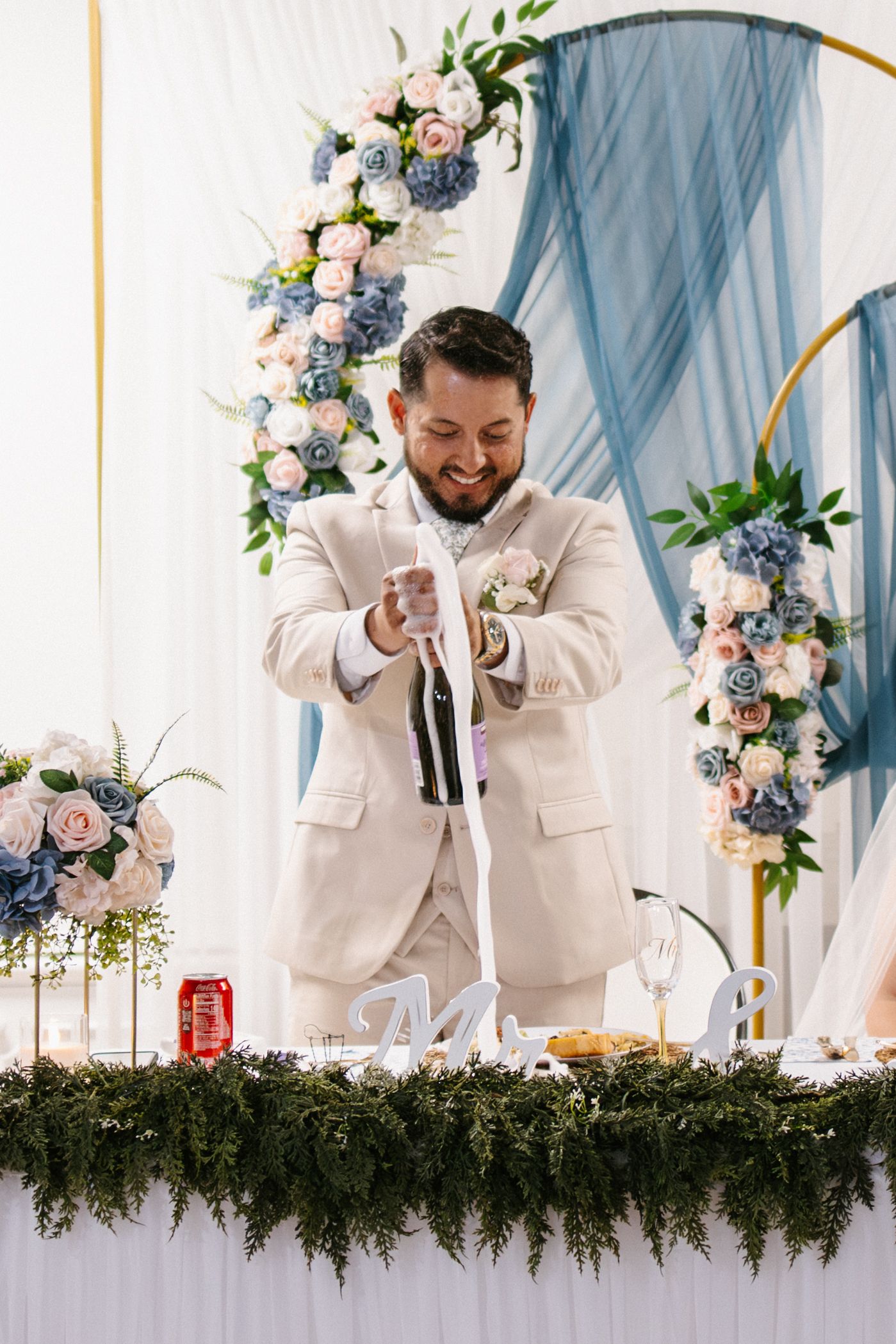 Smiling groom in beige suit opening champagne bottle at wedding reception table with blue and pink floral arch backdrop.