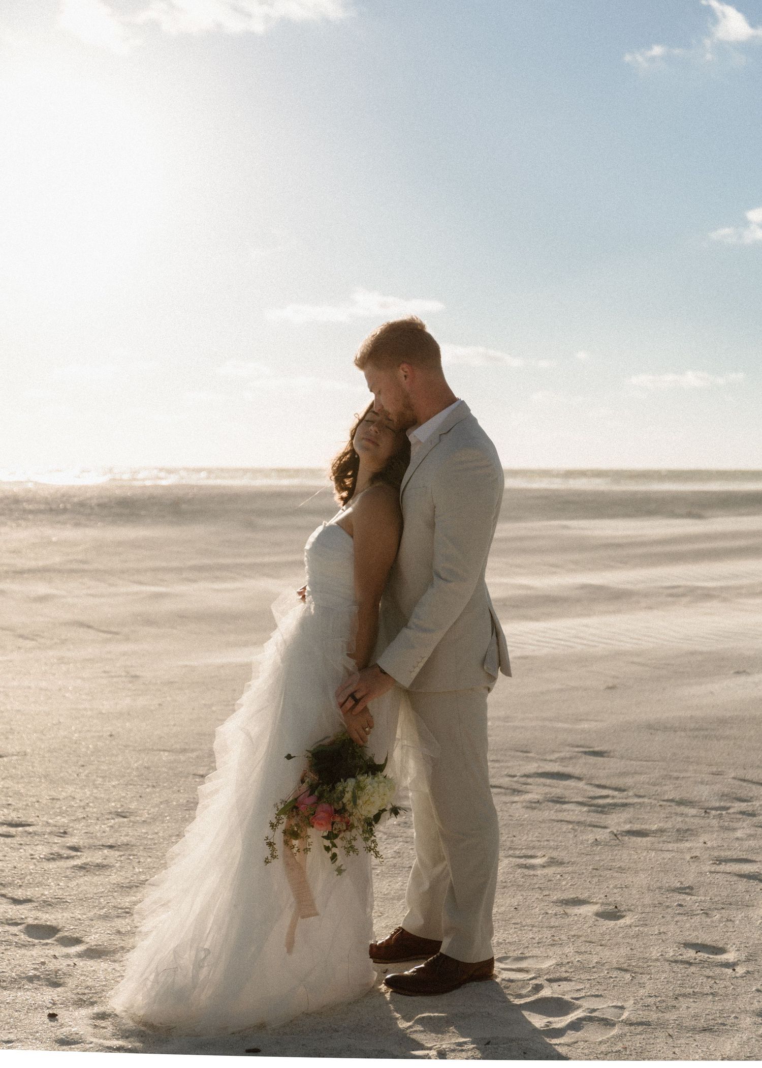 Bride and groom share a romantic embrace on a sunlit beach, with the bride holding a floral bouquet.