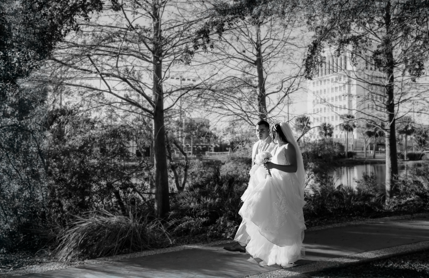 Black and white photo of a bride in a flowing gown posing outdoors near trees and a pond on her wedding day.