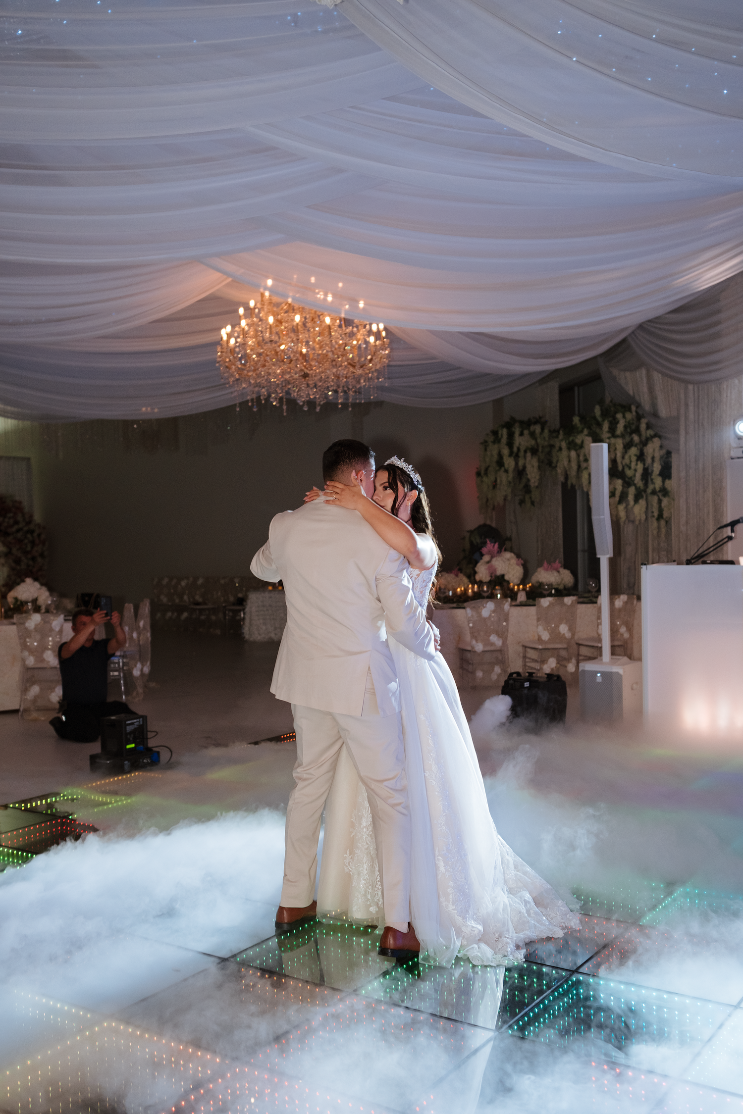 Newlyweds share their first dance on a mirrored floor surrounded by romantic fog and chandeliers.