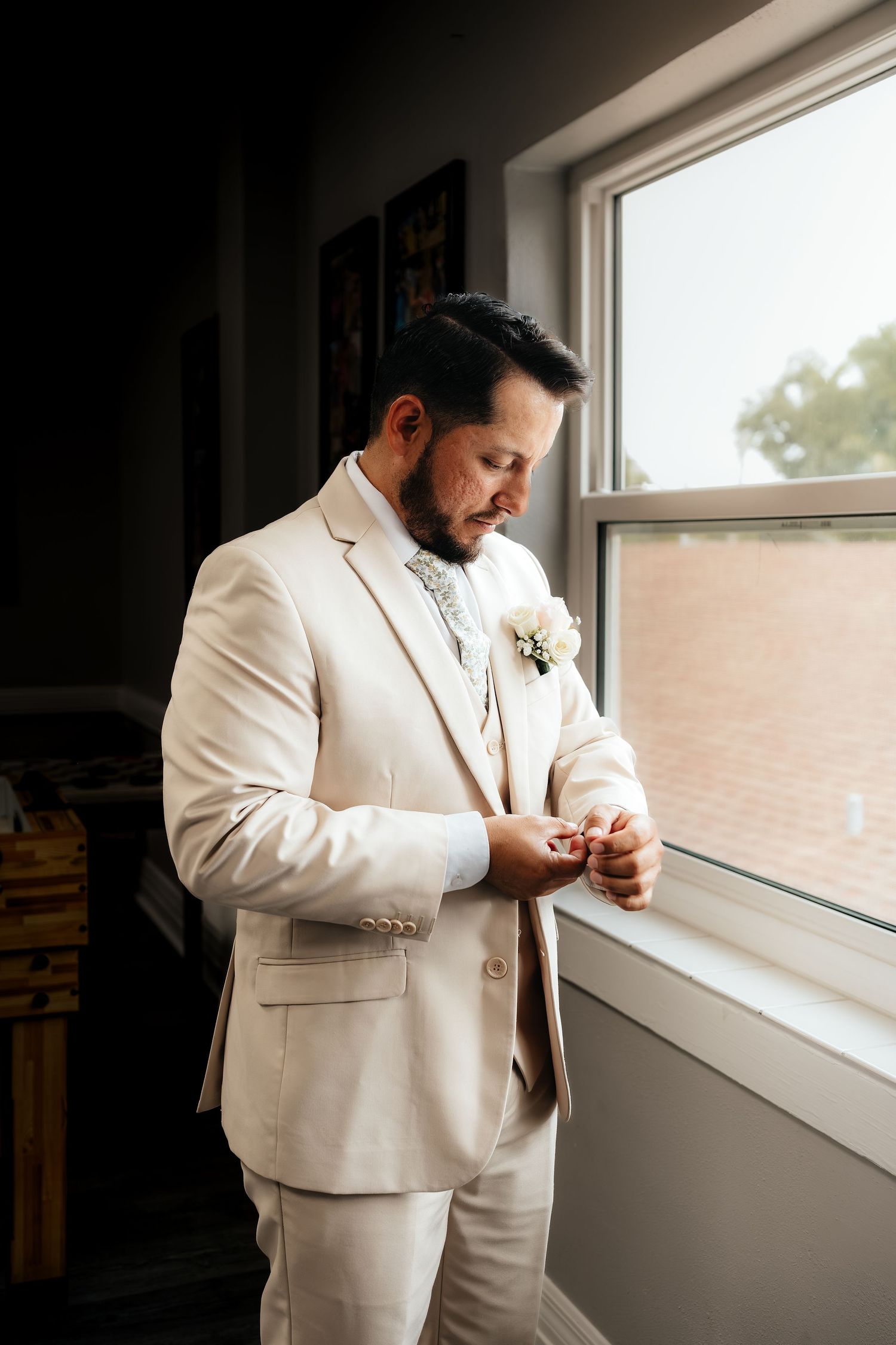 Groom in beige suit with white boutonniere adjusting cufflinks by window before wedding ceremony.