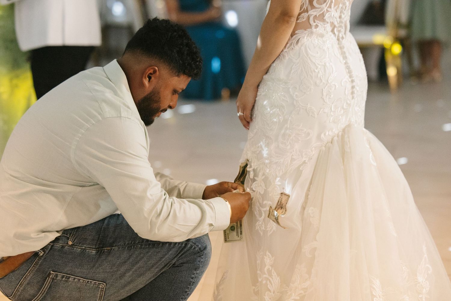 Groom kneels to pin money on brides lace mermaid wedding dress during festive dollar dance reception tradition.