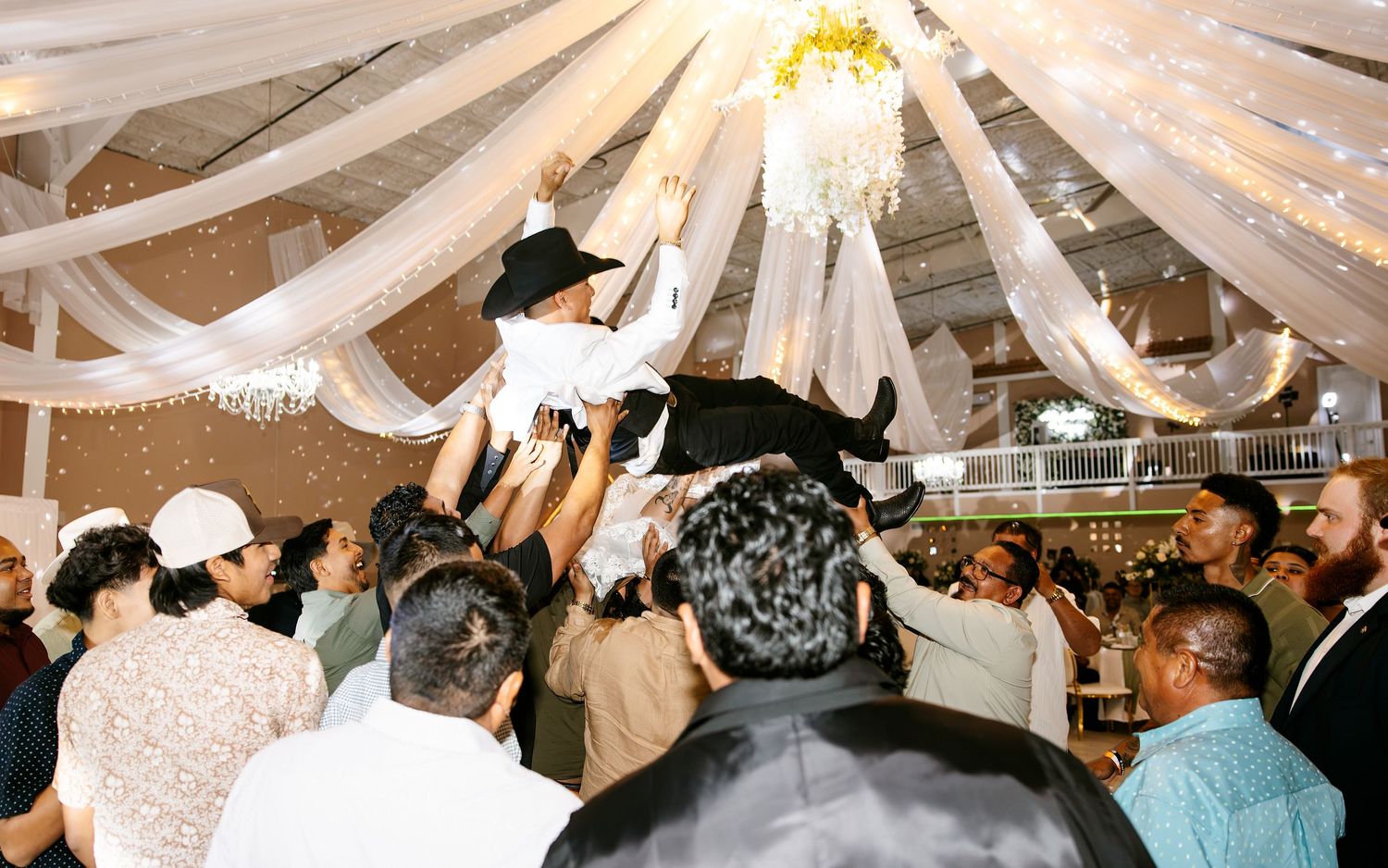 Guests lift a man in a cowboy hat during a festive wedding celebration under draped white fabric ceiling.