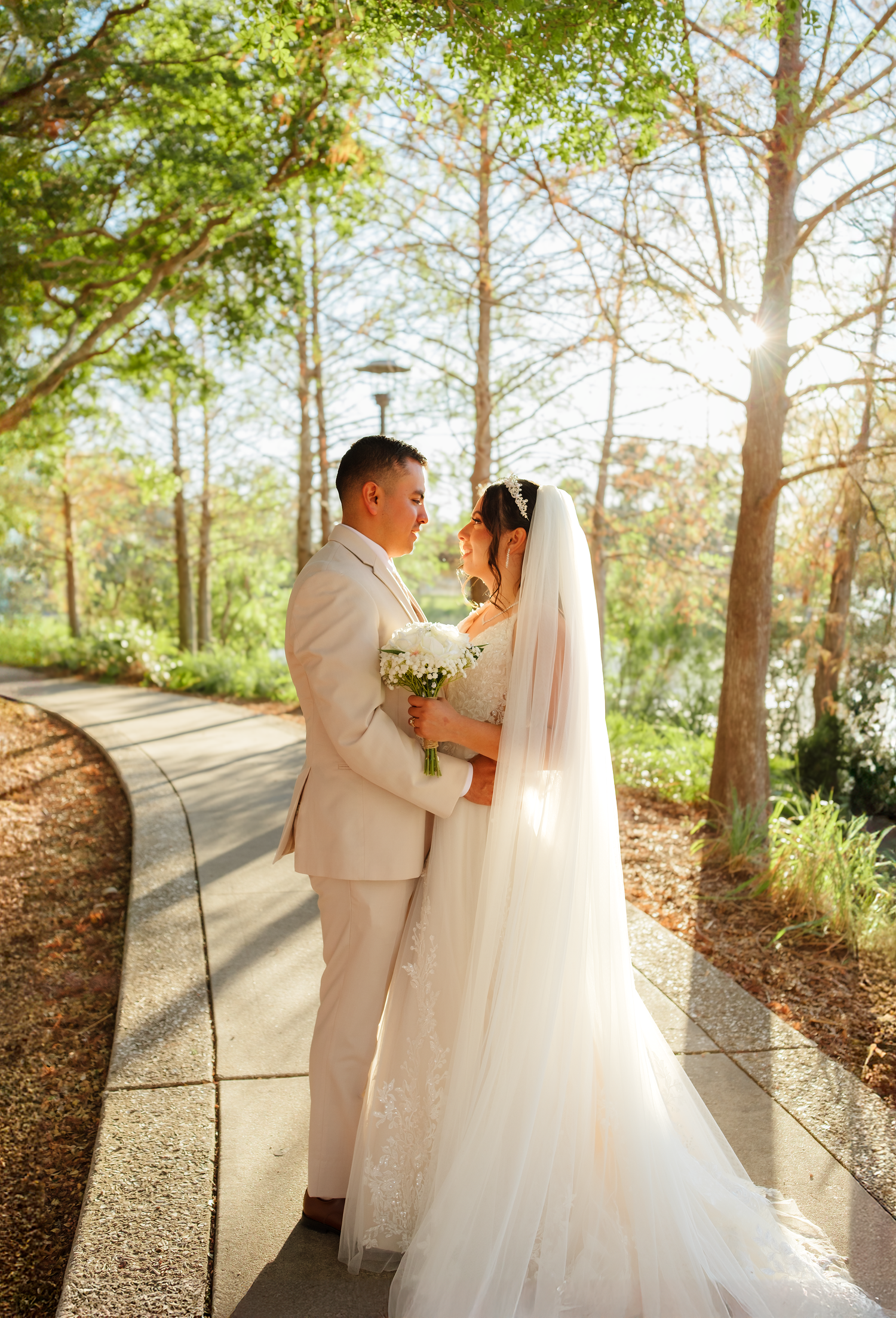Bride and groom share a romantic moment on a sunlit garden pathway surrounded by tall trees.