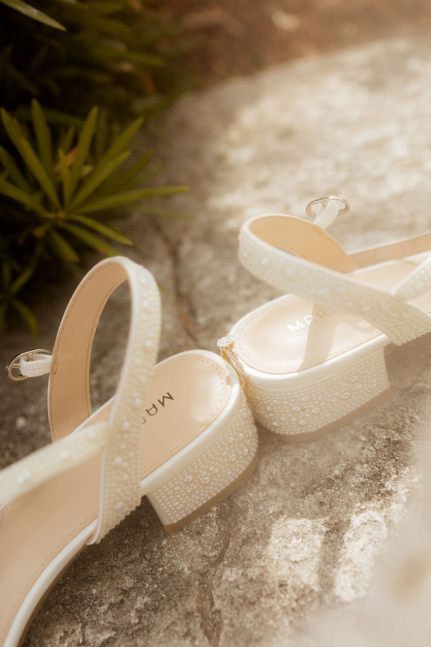 Pearl-embellished white bridal heels with ankle straps resting on stone surface in soft natural light.