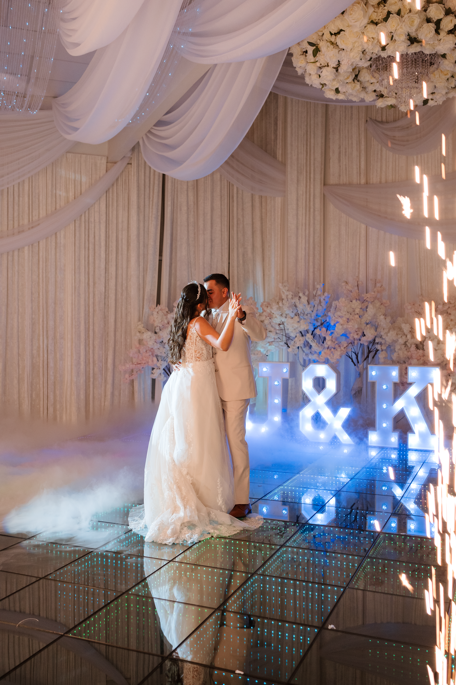 Newlyweds share their first dance on a mirrored floor beneath glowing J&K monogram letters.