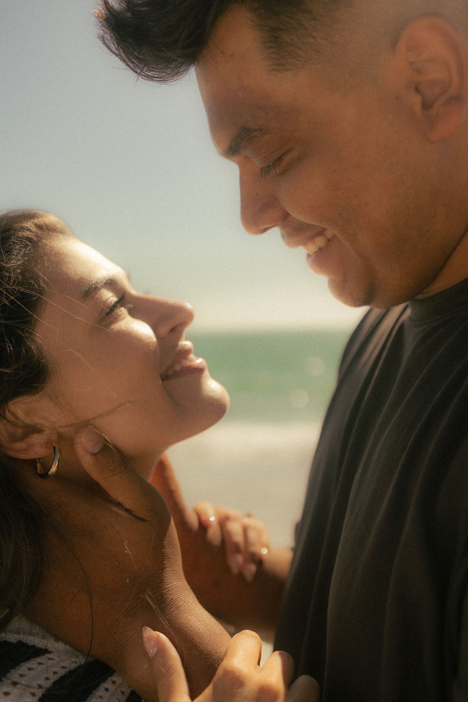 A smiling couple holds hands and gazes at each other intimately on a sunny beach with ocean waves in the background.
