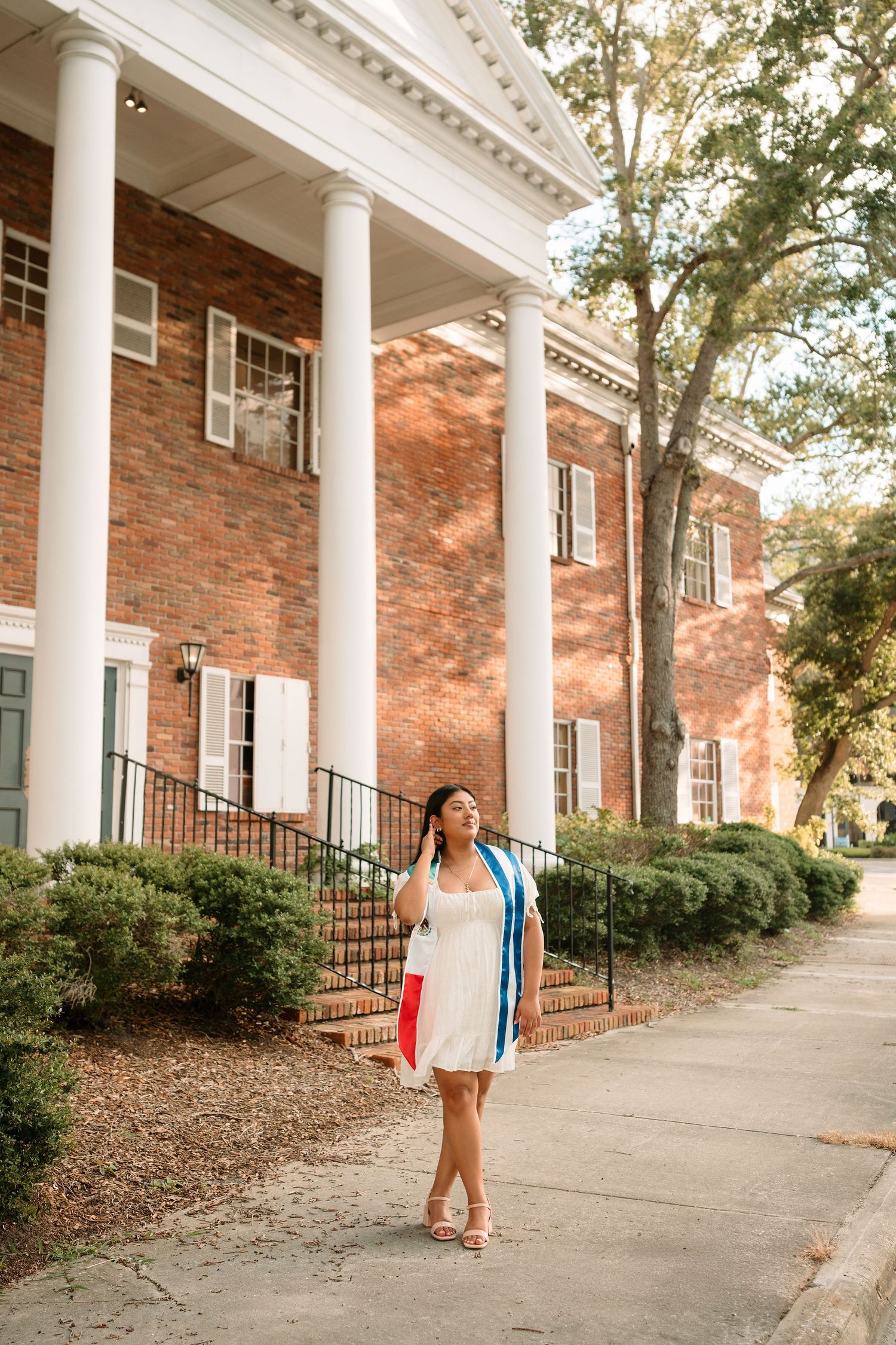 Young woman in white dress holding graduation regalia walks outside a brick building with white columns.