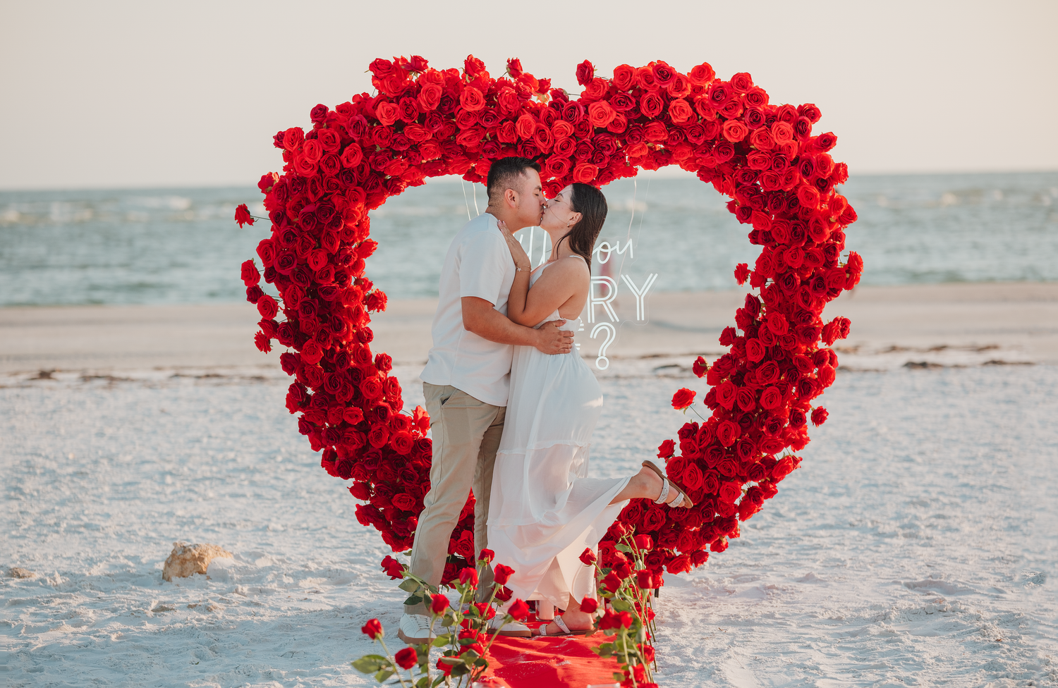 Couple kissing under a large heart-shaped red rose arch on a romantic beach at sunset.