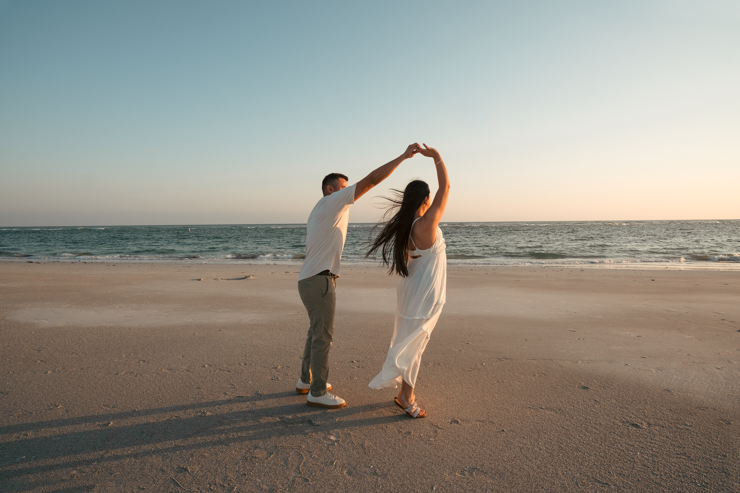 Couple dancing on a sandy beach at sunset, woman in white dress, man in white shirt, ocean in background.