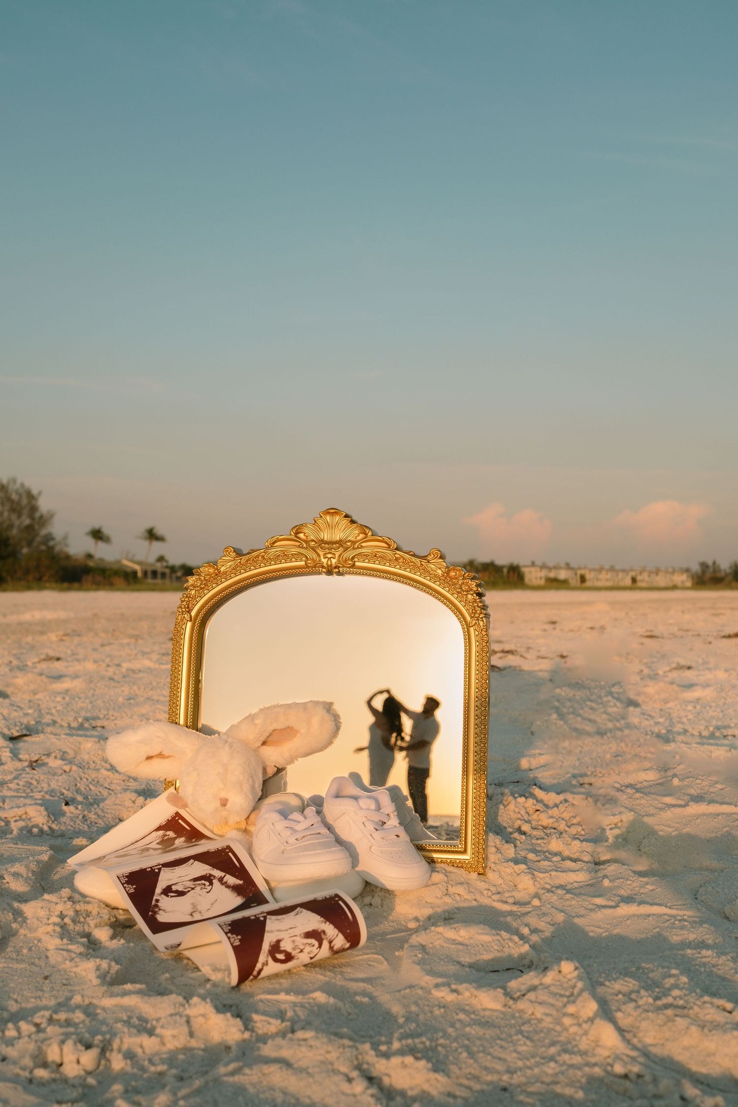 Gold ornate mirror on sandy beach reflects expecting couple, with baby shoes, ultrasound photos, and stuffed bunny in foreground.