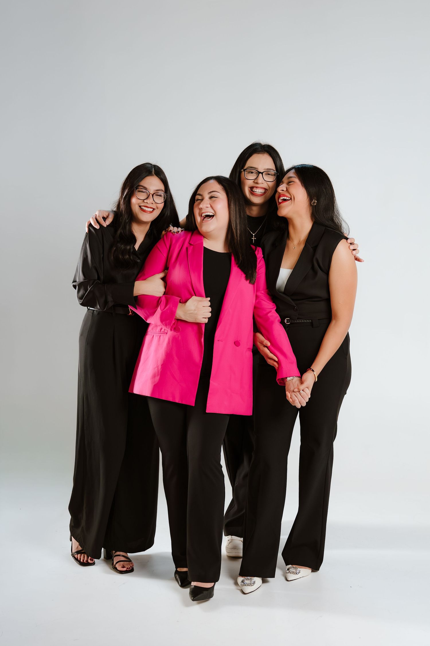 Four women in black outfits, one in pink blazer, laughing and embracing during a professional studio photo shoot.