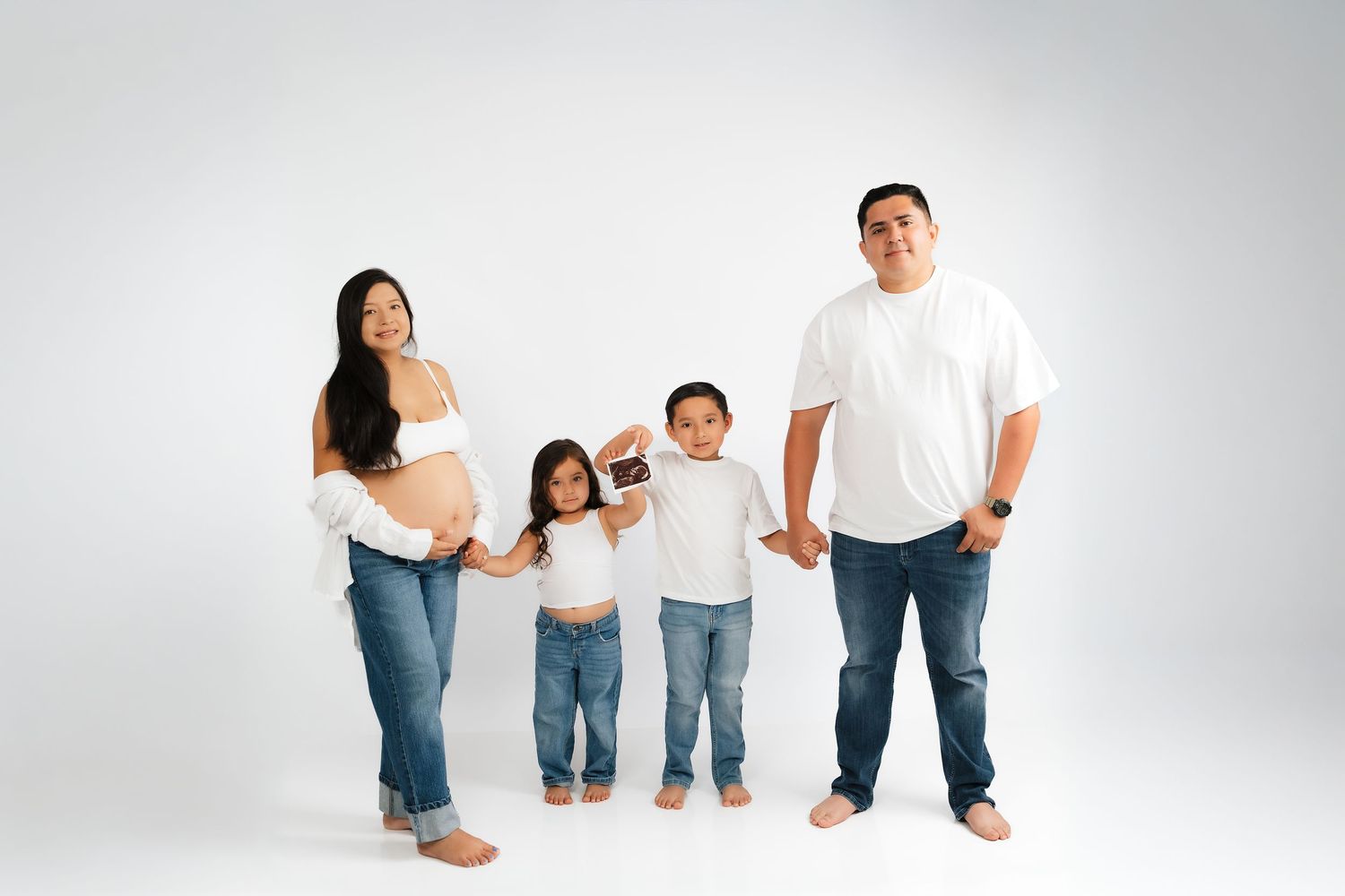 Pregnant Asian family of four posing together in white tops and jeans against a white studio background.