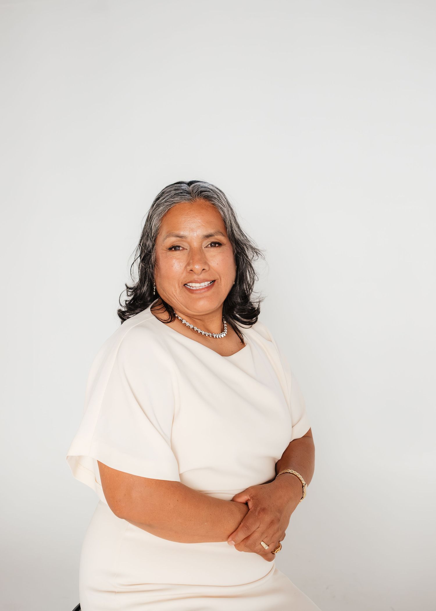 Professional woman in cream top with gray-streaked hair, arms crossed, smiling against white background.