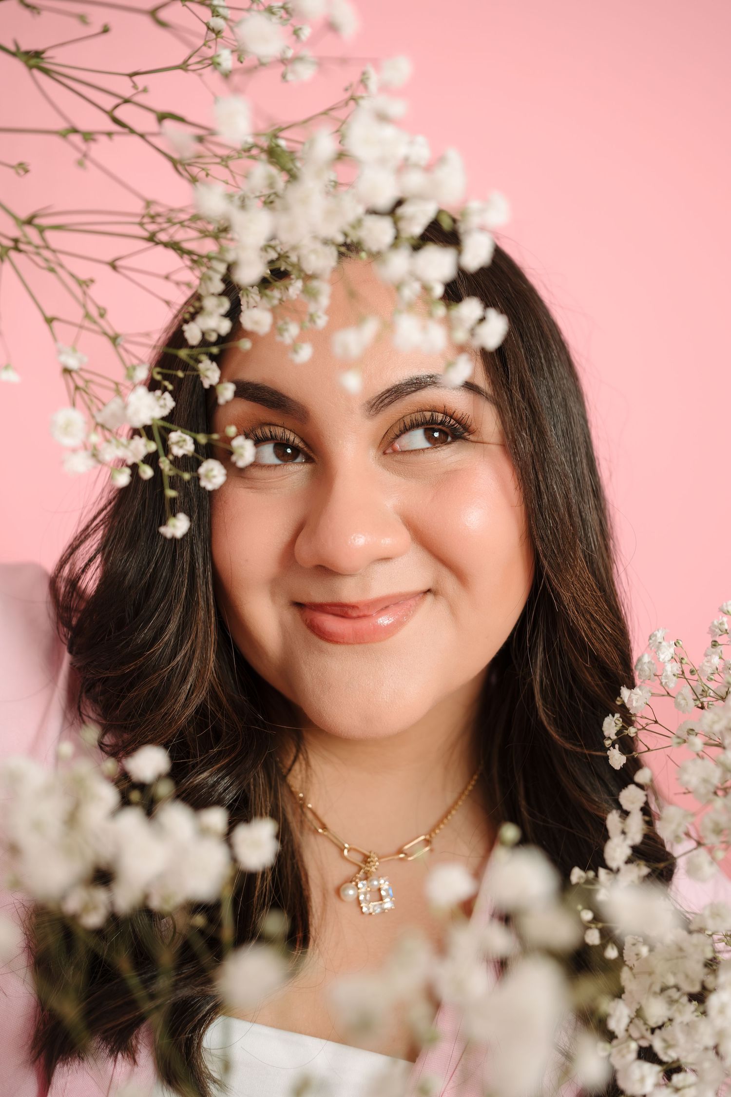 Young woman smiling surrounded by white baby's breath flowers against a pink background, wearing a gold necklace.