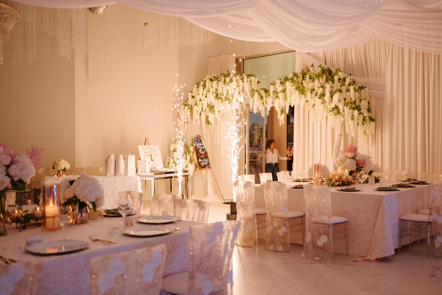 Elegant wedding reception hall with white floral ceiling installation, draped fabric, and gold candlelit table settings.