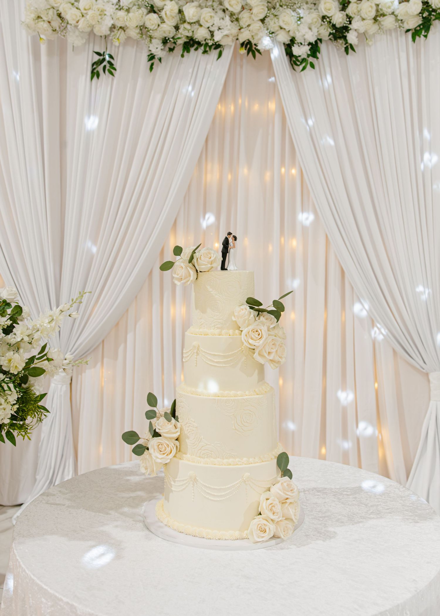 Elegant four-tier white wedding cake with rose decorations and bride and groom topper against draped white backdrop.
