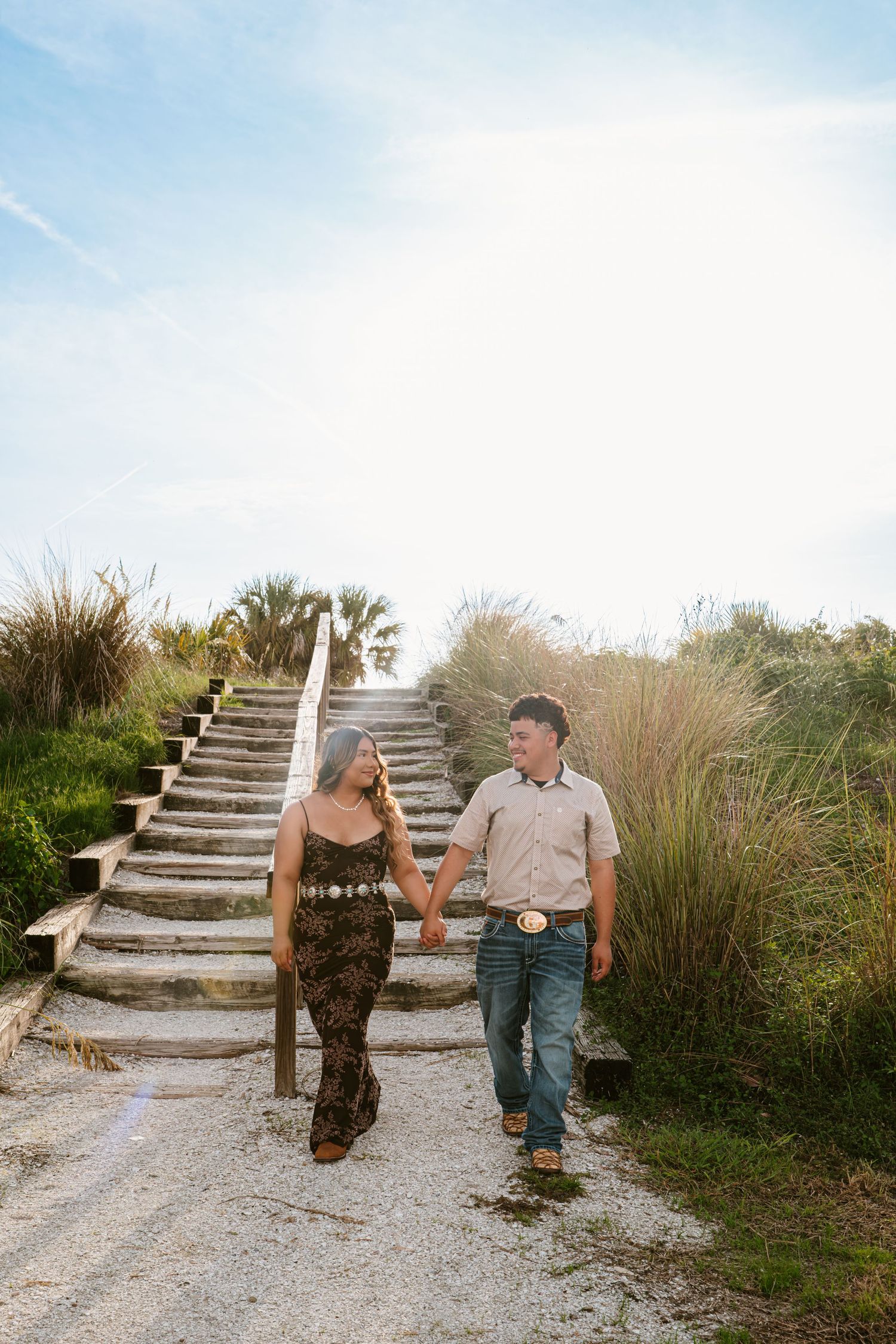 Couple holding hands walking down wooden beach stairs surrounded by tall grass and palm trees on a sunny day.