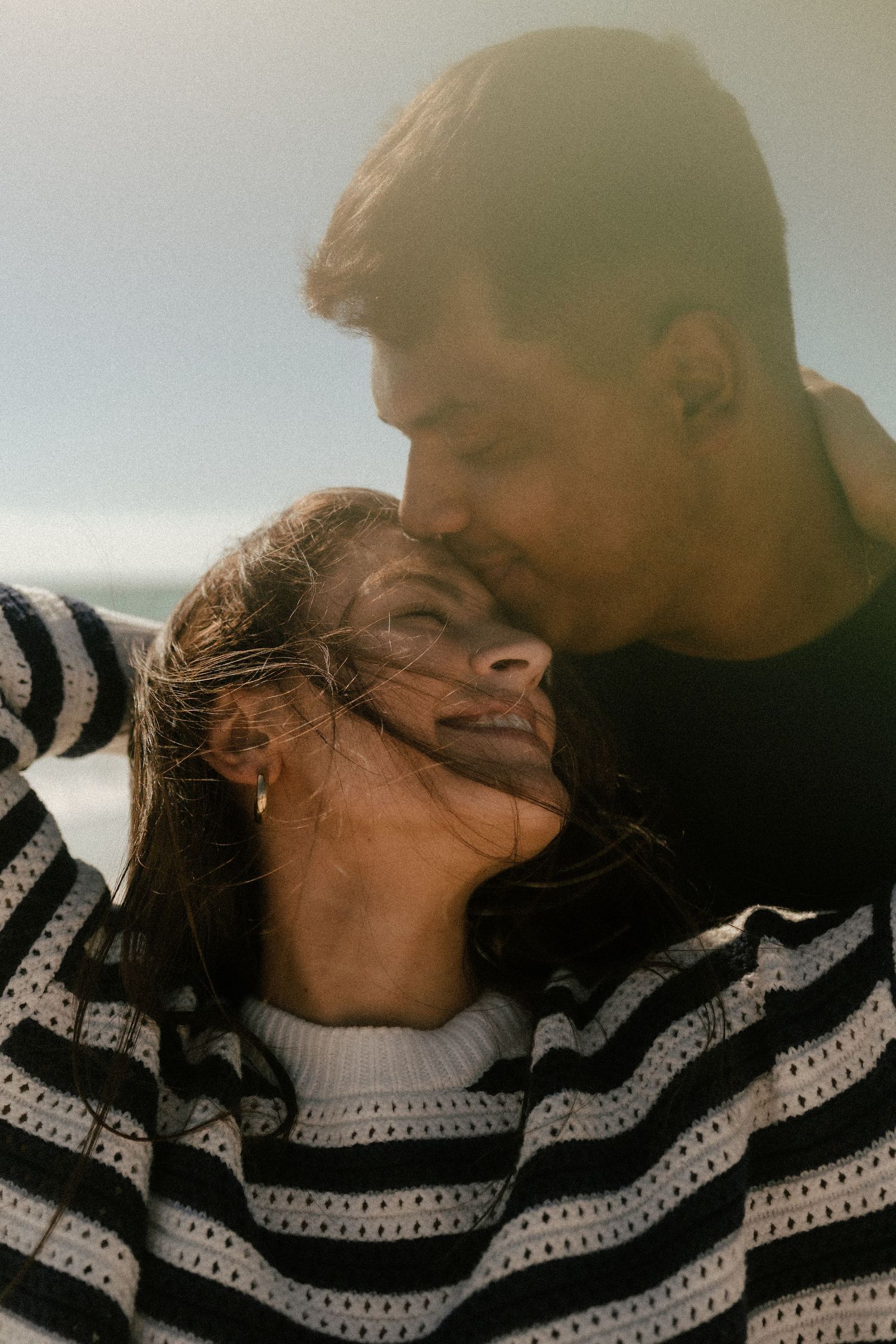 A couple shares a tender moment on a windy beach, with the man kissing the smiling woman on her forehead.