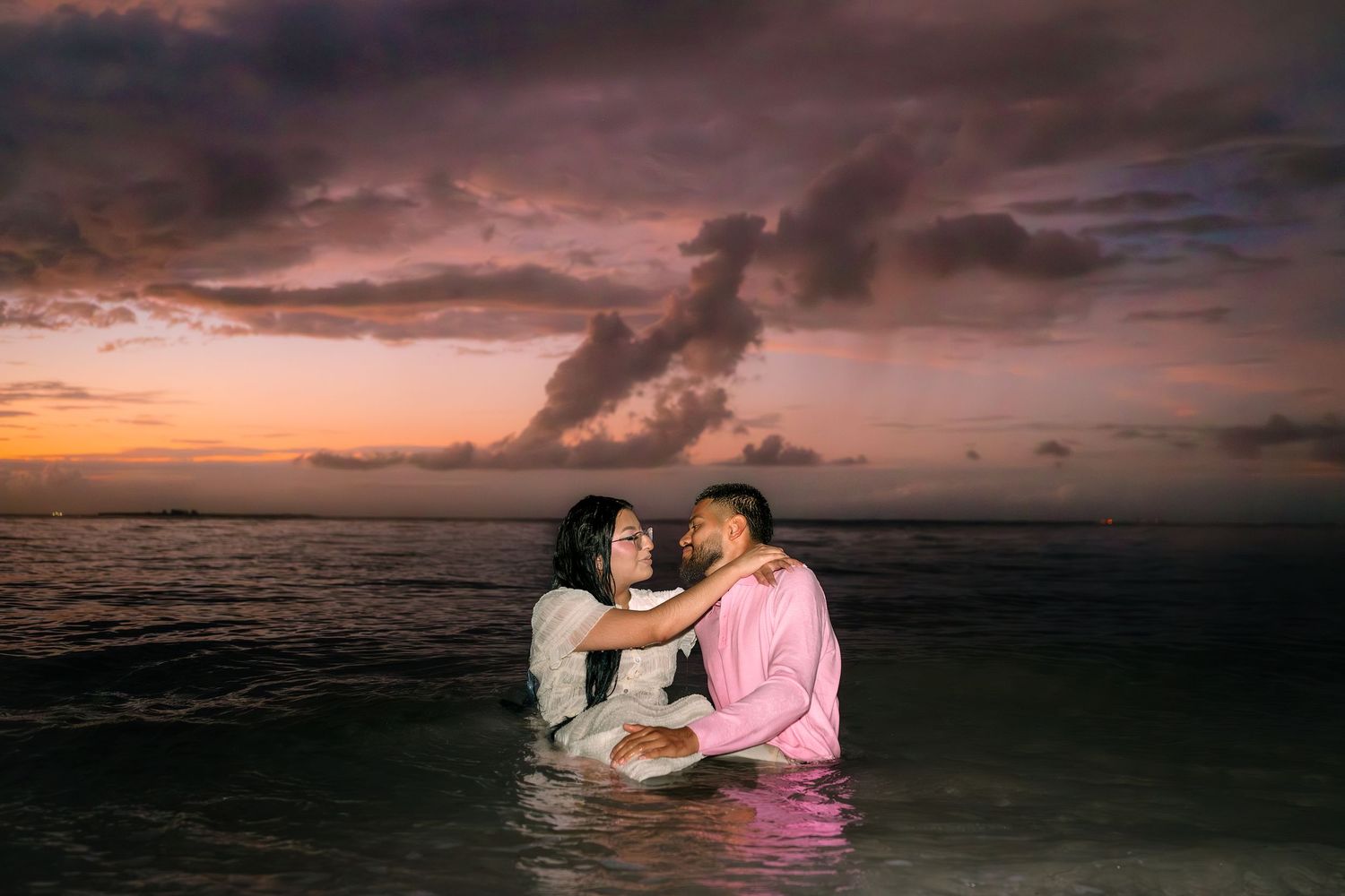 Couple embracing in ocean water at sunset with dramatic purple and pink cloudy sky backdrop.