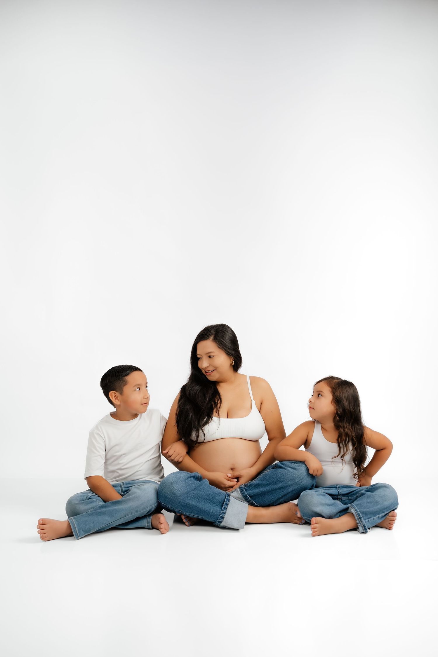 Pregnant woman sitting on white floor with two children, all wearing jeans and white tops, smiling together.